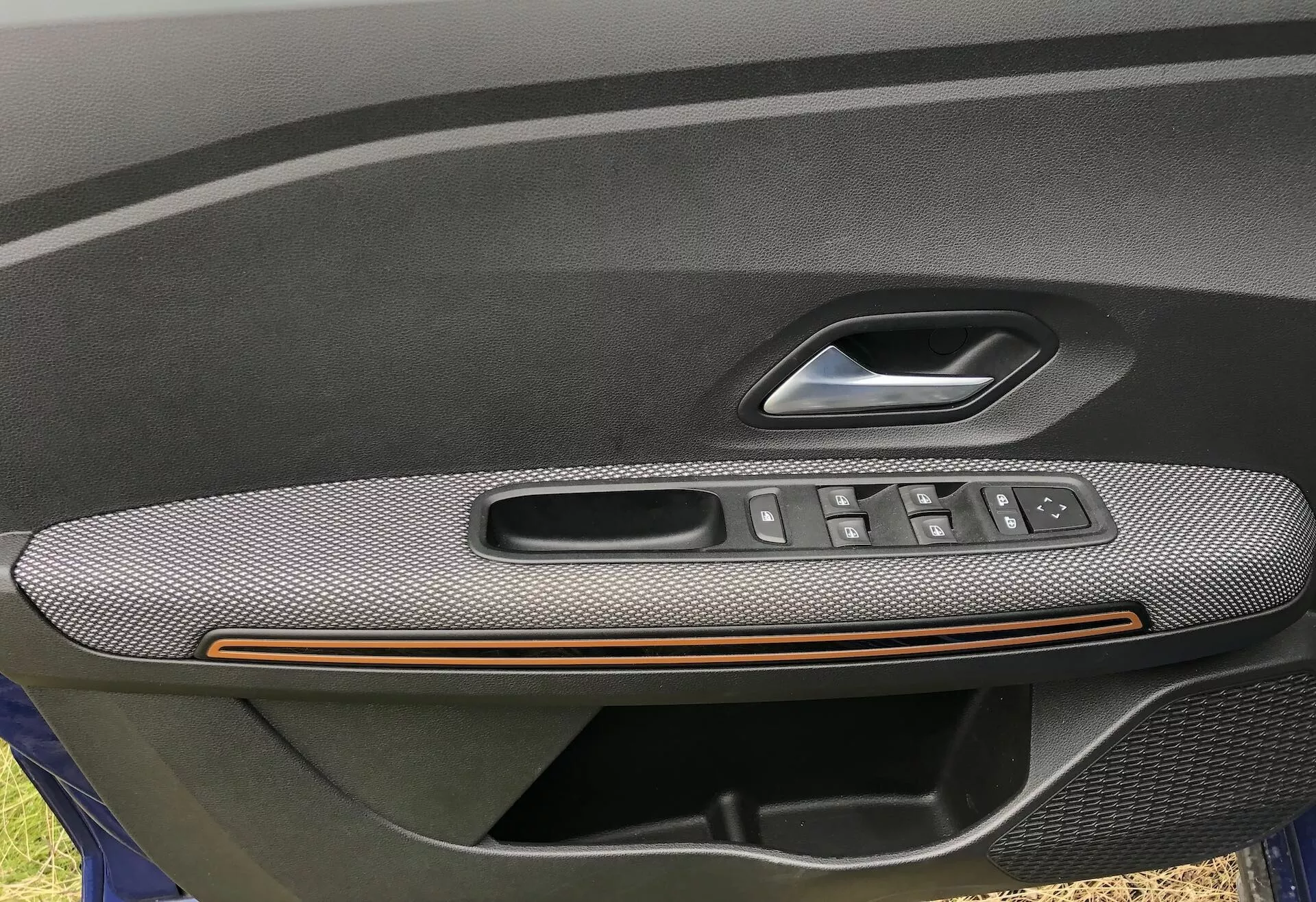Detailný záber na vnútorný panel dverí vodiča Dacia Sandero 2020 Stepway, ktorý zobrazuje opierku rúk s textúrou z uhlíkových vlákien, bronzovú akcentnú líniu a integrovaný ovládací panel s tlačidlami na elektrické okná, joystick na nastavenie bočných zrkadiel a kľučku dverí s tlačidlom zamykania/odomykania. Obrázok zdôrazňuje luxusné a praktické prvky, čo naznačuje vyššiu úroveň výbavy.