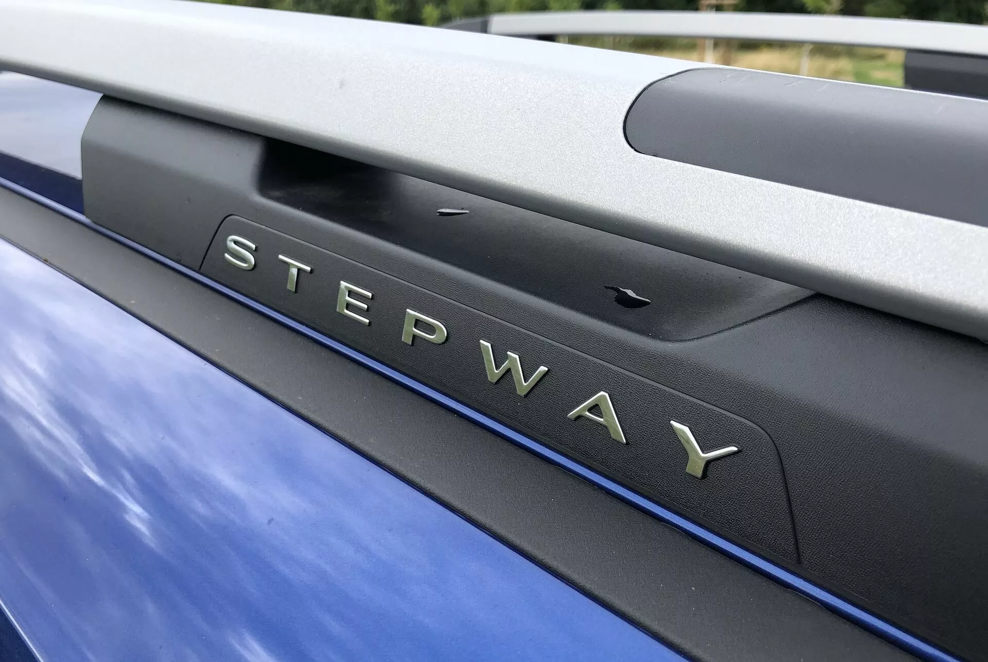Detailný záber na odznak 'STEPWAY' na Dacia Sandero 2020 (Sandero-Stepway) s metalickou modrou farbou. Odznak so striebornými písmenami s chrómovou úpravou je pripevnený na sekcii, ktorá pravdepodobne patrí k zadným dverám alebo kufru, doplnený čiernou lesklou lištou nad ním. Nad rámom fotografie je strešný nosič, ktorý naznačuje ďalšiu prepravnú kapacitu, ktorá zodpovedá dobrodružnému dizajnu vozidla. Vedľa odznaku je viditeľný parkovací senzor, čo naznačuje moderné estetické a bezpečnostné prvky.
