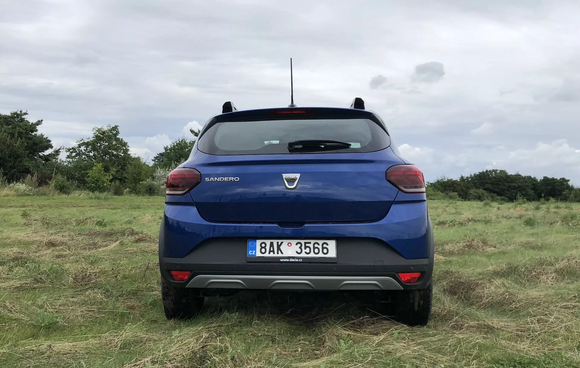 Zadný pohľad na modrú Daciu Sandero Stepway 2020 zaparkovanú na nerovnom trávnatom poli, so značkou a názvom modelu výrazne zobrazenými na zadných dverách. Zadné sklo vozidla má jediný stierač, na bokoch sú viditeľné zadné svetlá s červeno-bielou farebnou schémou. Viditeľné sú vysoké brzdové svetlo pri základni zadného okna, strešná anténa a dvojfarebný zadný difúzor. Nárazník má možné parkovacie senzory, ŠPZ je centrálne pod logom značky a prehlbina môže slúžiť ako priestor pre kameru.