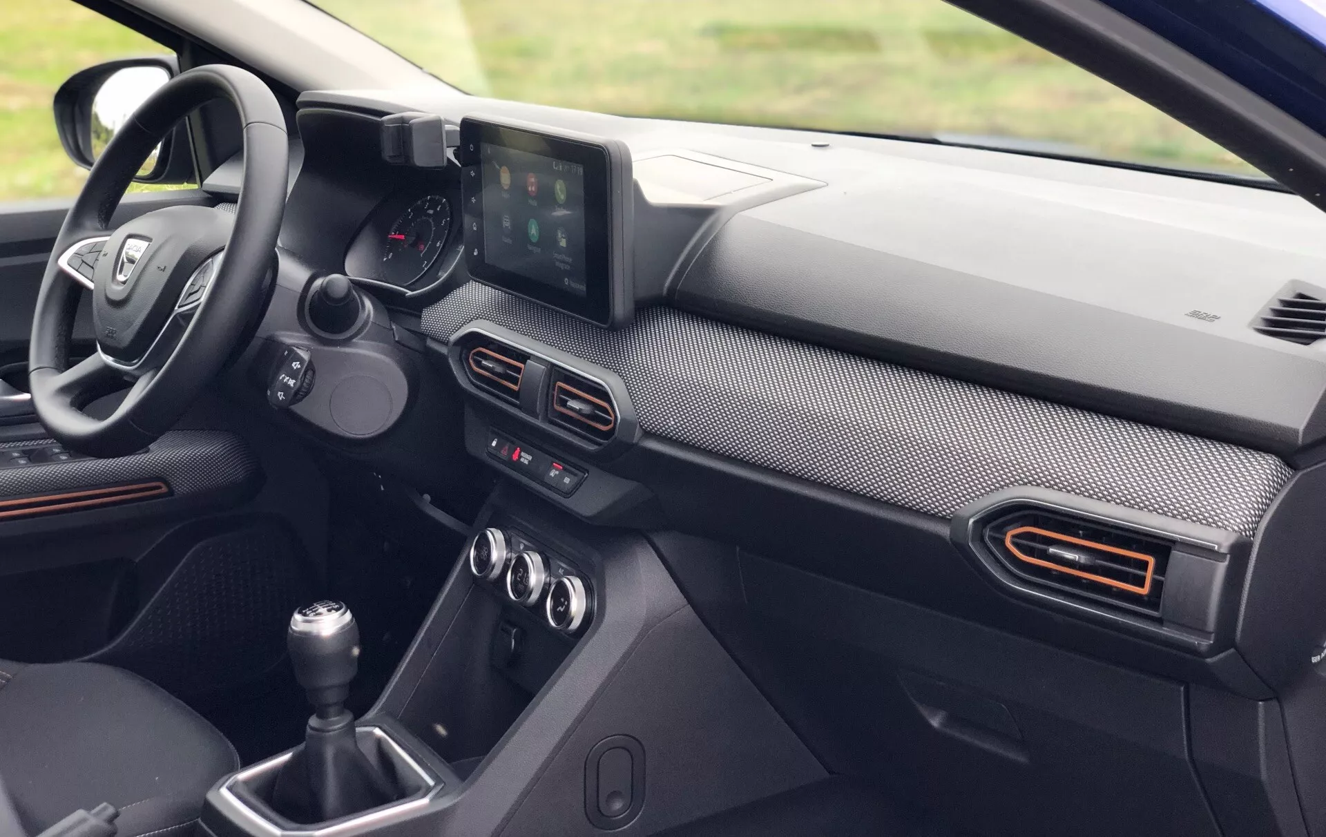 Detailný pohľad na interiér Dacia Sandero Stepway 2020, zobrazujúci prístrojovú dosku na strane vodiča z perspektívy predného spolujazdca. Viditeľné prvky zahŕňajú multifunkčný volant s ovládacími tlačidlami, kombinovaný analógový a digitálny prístrojový panel, samostatný infotainment displej a ručnú radiacu páku. Prístrojová doska má povrchovú úpravu podobnú uhlíkovým vláknam a možné farebné akcenty okolo vetracích otvorov, čo naznačuje moderný a možno výkonný dizajn.