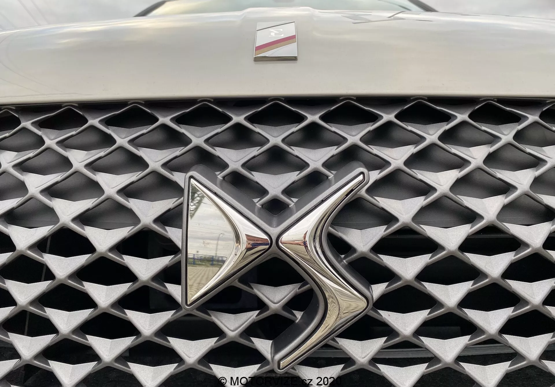 Detailný záber na mriežku modelu DS Automobiles DS 3 Crossback E-Tense 2019. Obrázok zachytáva mriežku z čelného pohľadu, pričom sa zameriava na výrazný, vysoko reflexný kovový znak. Mriežka má charakteristickú textúru tvorenú diamantovými bunkami v matnej sivej farbe, ktorá kontrastuje s lesklým chrómovým znakom. Obmedzené zorné pole mimo mriežku a znak, rozmazané pozadie.