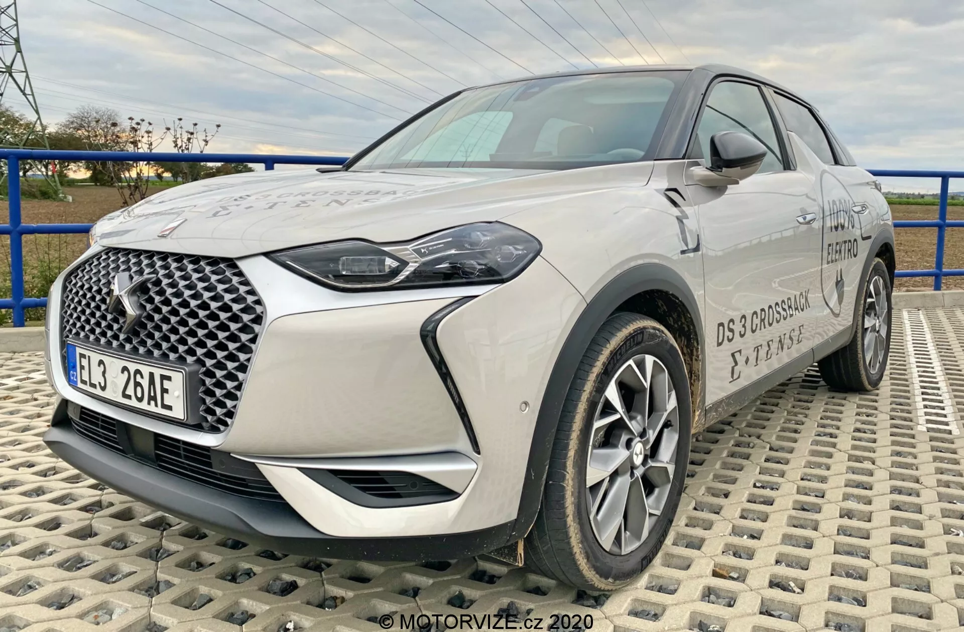 Obrázok ukazuje moderné crossover SUV DS-Automobiles DS-3 2019 (DS-3 Crossback E-Tense), zachytené z uhla troch štvrtín spredu na textúrovanej ploche tvořenej prepletenými dlaždicami. Perspektíva z nízkej až strednej výšky zdôrazňuje postavu auta a poskytuje komplexný pohľad na jeho prednú mriežku s výrazným geometrickým vzorom, LED svetlami, hliníkovými kolesami, zrkadlami a detailmi značky. Dĺžka a predné dizajnové prvky vozidla sú jasne viditeľné, čo poukazuje na jeho modernú a pokročilú automobilovú technológiu.
