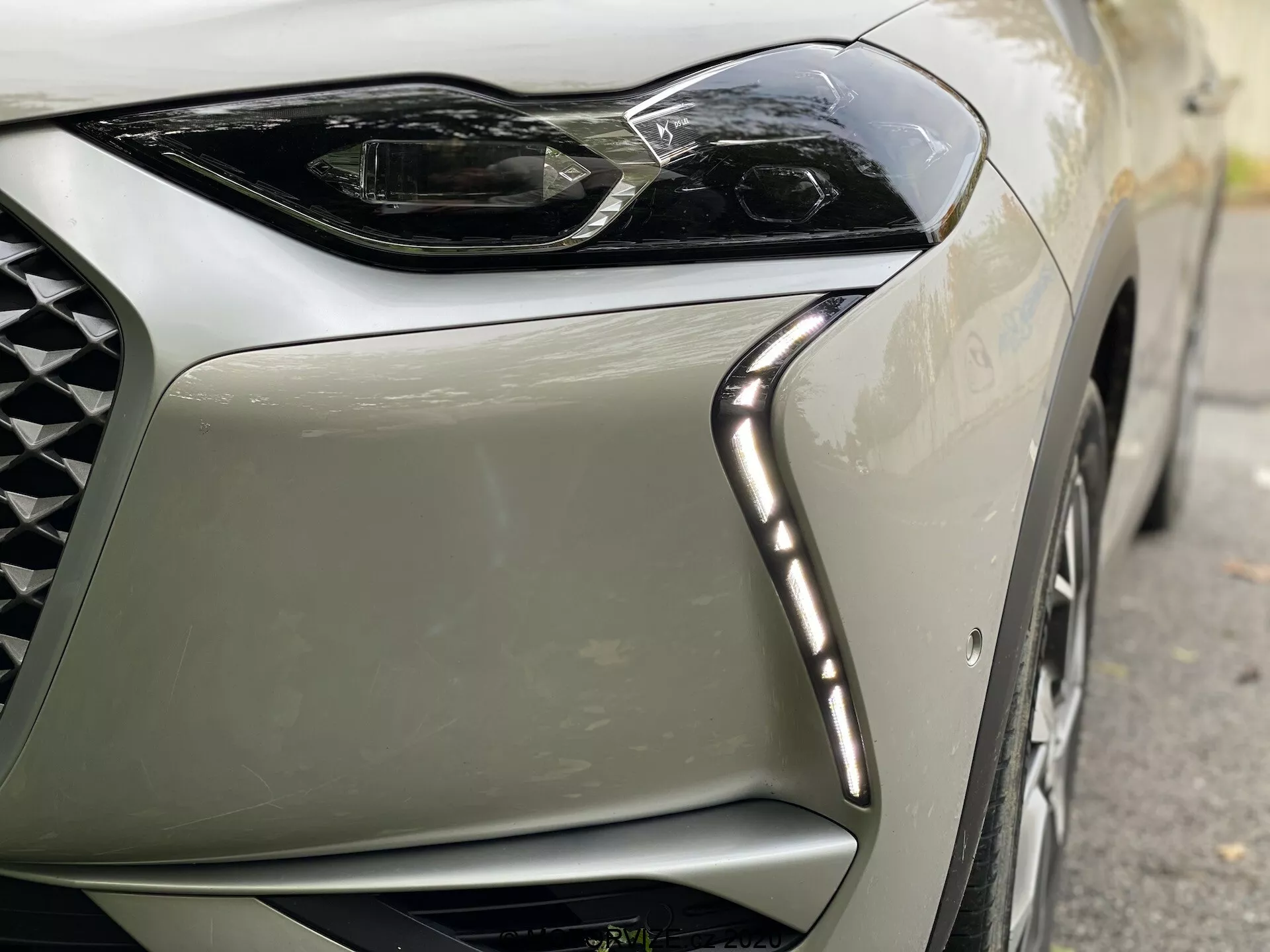 Detailný pohľad na predný ľavý roh moderného vozidla DS Automobiles DS-3 Crossback E-Tense z roku 2019. Obrázok sa zameriava na elegantný, uhlový dizajn LED svetiel s LED dennými svetlami pozdĺž horného okraja a časť mriežky s diamantovým vzorom, zdôrazňujúci luxus a špičkovú technológiu.
