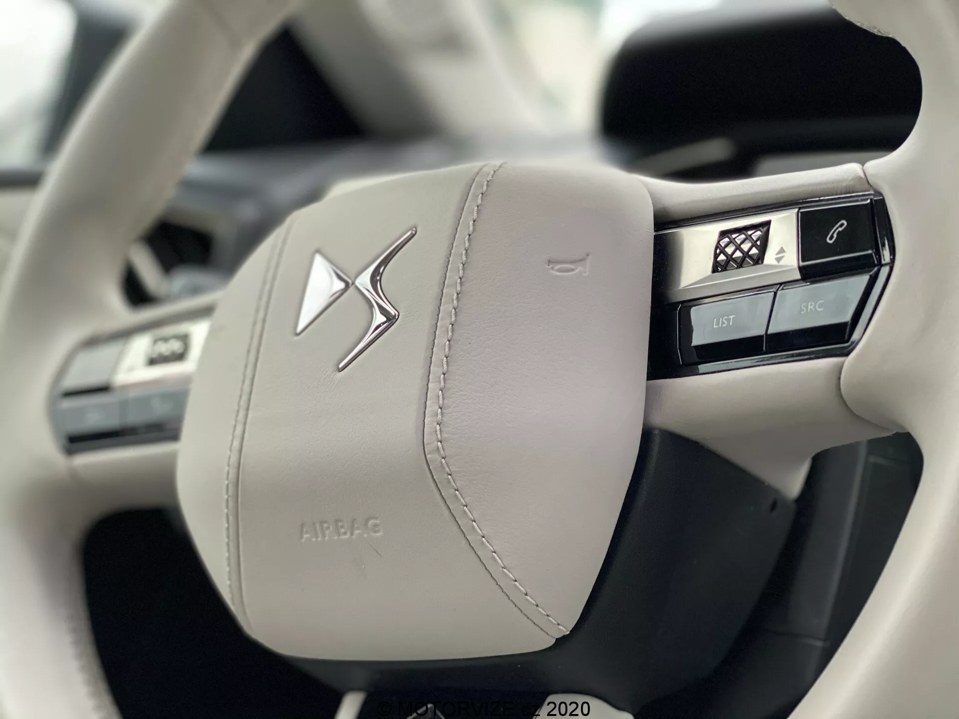 Detailný pohľad na ľavú stranu moderného, svetlo sfarbeného koženého volantu vozidla DS Automobiles DS 3 Crossback E-Tense 2019, zdôrazňujúci remeselnú výrobu a luxus s viditeľným šitím. Obsahuje niekoľko integrovaných tlačidiel pre ovládanie funkcií vozidla, vrátane hlasového ovládania, telefónu, hlavného menu a ovládania médií, vedľa výrazného centrálneho emblému a púzdra s nápisom 'AIRBAG'.