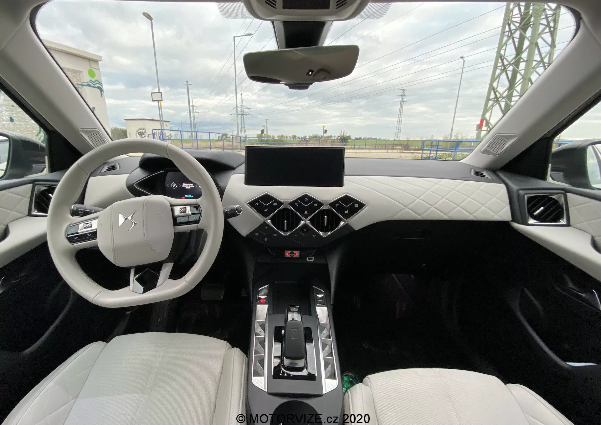 Pohľad zo zadného sedadla na moderný centrálny interiér vozidla DS Automobiles DS-3 Crossback E-Tense 2019. Fotografia zachytáva trojramenný volant s ovládacími prvkami, digitálny displej prístrojového panela, veľkú dotykovú obrazovku na stredovej konzole s hexagonálnymi tlačidlami klimatizácie pod ňou a radiacu páku naznačujúcu automatickú prevodovku. Palubná doska a dvere majú prešívaný dizajn z koženky alebo kože, predné športové sedadlá sú čalúnené svetlou kožou alebo syntetickým materiálom. Viditeľné sú aj ambientné osvetlenie pozdĺž obloženia, panoramatická strecha a výduchy vzduchu na palubnej doske.