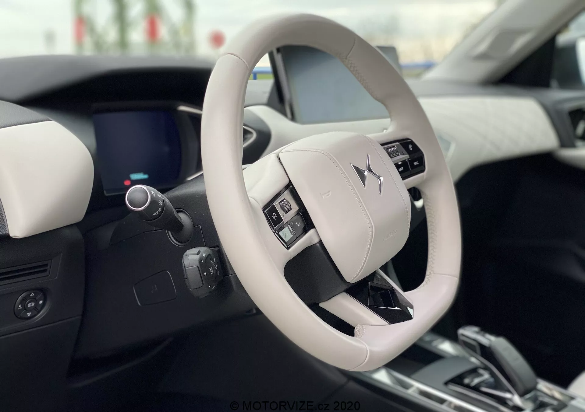 Vnútorný pohľad na DS Automobiles DS 3 Crossback E-Tense 2019, zobrazujúci moderný a luxusný dizajn volantu a palubnej dosky z perspektívy sedadla vodiča. Svetlo zafarbený volant obsahuje ovládacie tlačidlá a zreteľne zobrazuje logo výrobcu. Elegantná digitálna palubná doska, čiastočne viditeľná za volantom, dopĺňa dvojtónový interiér vysoko kvalitnými materiálmi.
