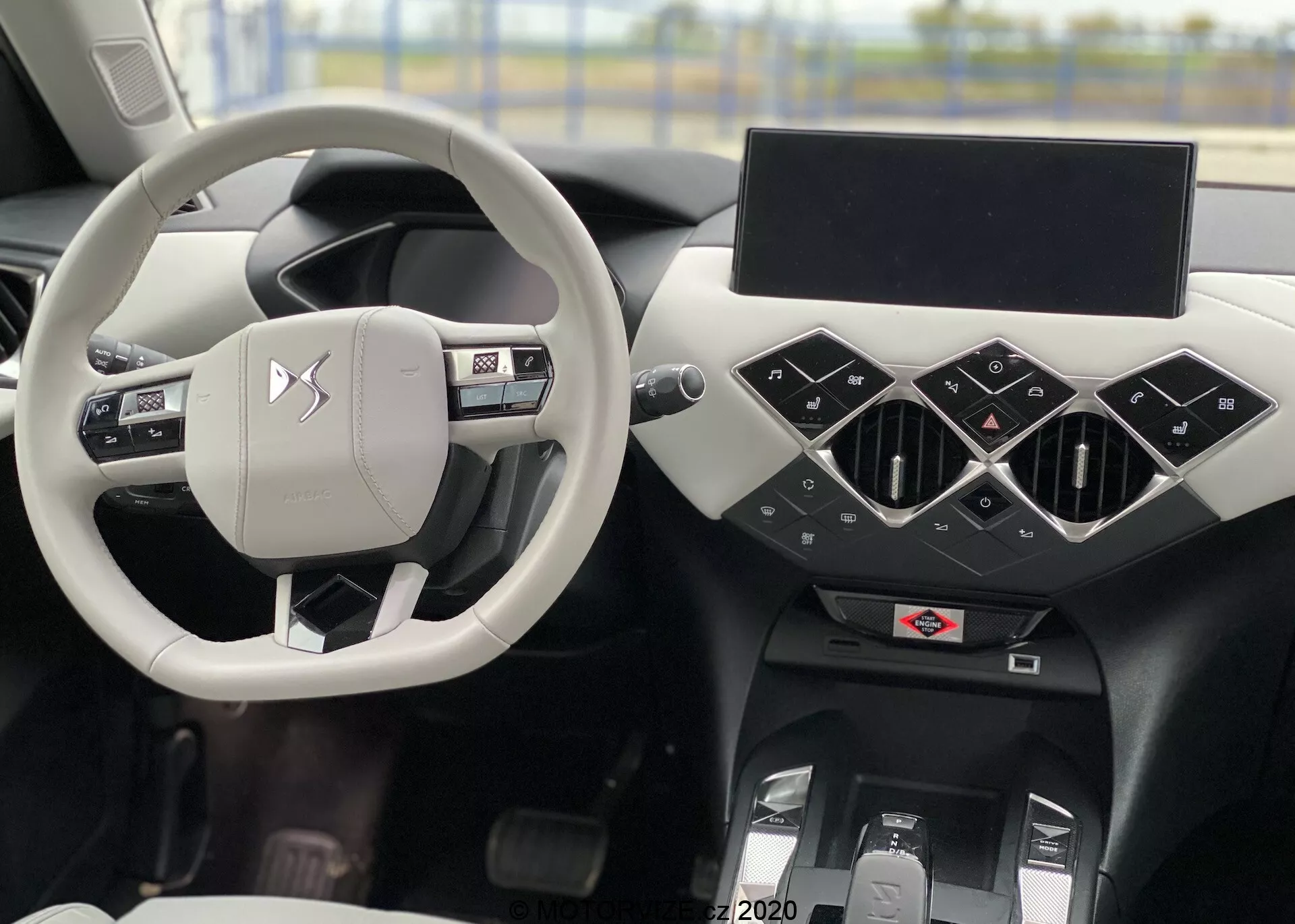 Interiérový záber modelu DS Automobiles DS 3 Crossback E-Tense z roku 2019 s dominantným strieborným/svetlosivým volantom s ovládacími tlačidlami a logom výrobcu, cez ktorý je vidieť sedadlo vodiča. Čierna palubná doska nesie geometrické prvky s trojuholníkovými stredovými ventilátormi, vypnutý infotainment displej a nižšie možné ovládacie prvky klimatizácie a centrálny tunel. Vľavo je vidieť časť dverí vodiča s ovládaním okien a zrkadiel.