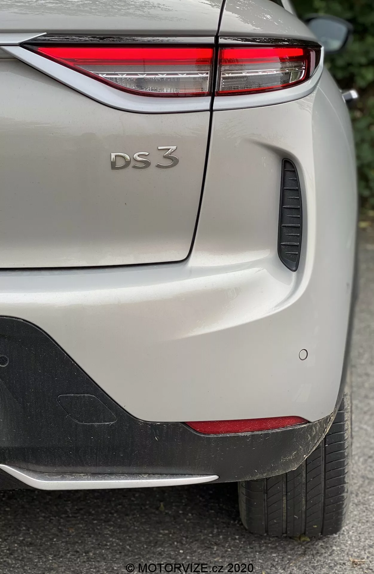 Detailný záber zadnej bočnej časti DS Automobiles DS 3 Crossback E-Tense 2019, zameriavajúci sa na hornú pravú časť zadného nárazníka, veko batožinového priestoru a časť pravého zadného blatníka. Obrázok zachytáva charakteristické dizajnové prvky modelu, vrátane tenkých zadných LED svetiel, reliéfneho označenia 'DS 3', parkovacích senzorov a čierneho plastového prvku na nárazníku. Auto je zobrazené v svetlom odtieni s metalickým povrchom a je fotografované na dennom svetle, čo zdôrazňuje jemné detaily laku a textúry.