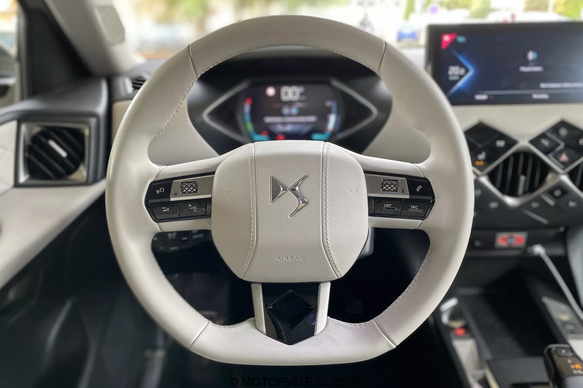 Pohľad z interiéru z perspektívy vodiča na DS Automobiles DS 3 Crossback E-Tense 2019, so zameraním na moderný volant s dvoma ramenami, pokrytý svetlou kožou, s multifunkčnými ovládacími prvkami a digitálnym prístrojovým panelom zobrazujúcim rýchlosť a otáčky. Stredová konzola obsahuje integrovanú dotykovú obrazovku infotainmentu a minimalistické ovládacie prvky klimatizácie, čo zdôrazňuje pokročilé automobilové technológie a luxusný dizajn.