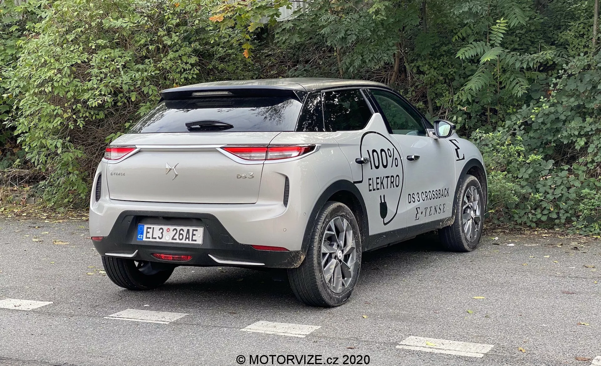 Automobil DS Automobiles DS 3 2019 (DS 3 Crossback E-Tense) zachytený z uhla zozadu troch štvrtín, zdôrazňujúci zadný a bočný profil vozidla. Dizajnové prvky zahŕňajú horizontálne orientované zadné svetlá, chrómovú lištu, zapustené kľučky dverí, nabíjací port a viacramenné zliatinové kolesá, ktoré sú typické pre moderný elektromobil. Auto je zaparkované vonku v mestskom alebo predmestskom prostredí pod zamračenou oblohou, s listnatými stromami neďaleko.