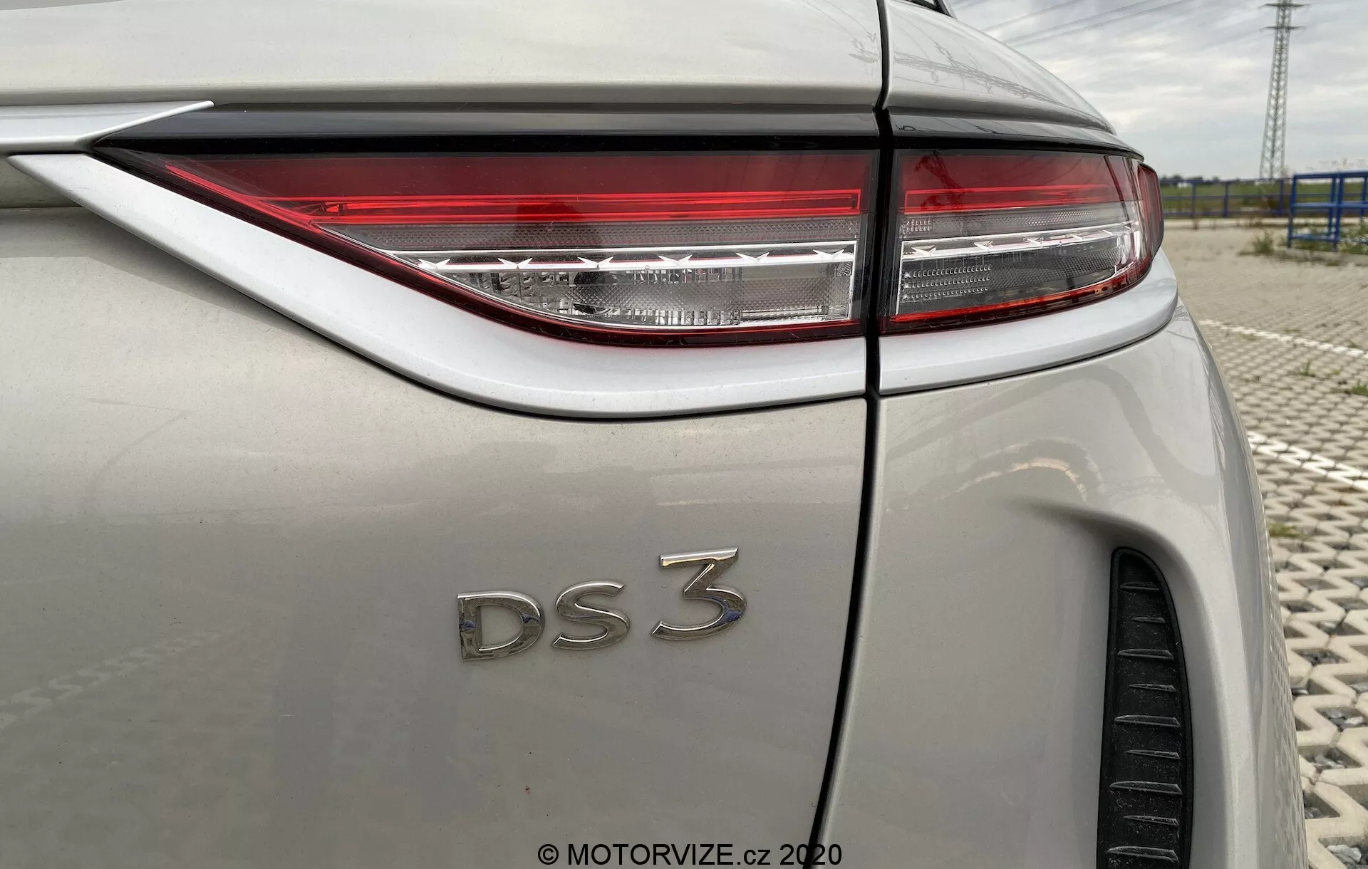 Zblízka zadného trojštvrťového uhla DS Automobiles DS 3 Crossback E-Tense z roku 2019. Obrázok sa zameriava na pravú stranu vozidla, zvýrazňujúc ostré LED zadné svetlá, odznak 'DS 3' a svetlú metalickú karosériu. Viditeľné prvky zahŕňajú líniu záhybu zadného blatníka, čiastočne viditeľnú zadnú pneumatiku, anténu typu žraločia plutva a prechod zadného okna do línie strechy, všetko pod odrazom oblačnej oblohy.