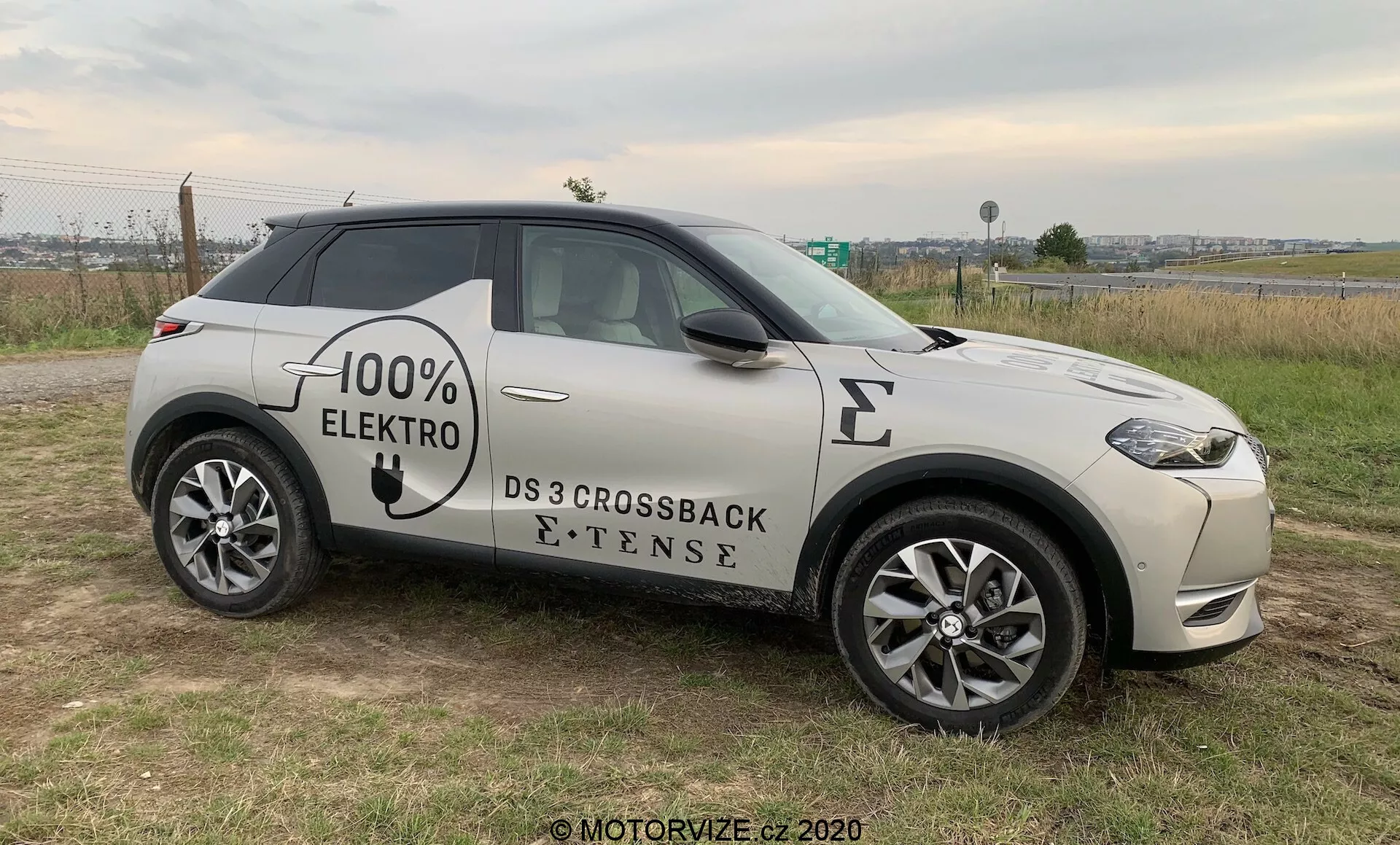 Bočný profil moderného SUV, DS 3 Crossback E-Tense z roku 2019, elektrického vozidla s grafickým nápisom '100% ELEKTRO' na boku. Automobil je umiestnený na trávnatom povrchu s kúskom spevnenej cesty v pozadí, čo umožňuje jasný pohľad na prednú mriežku, LED svetlomety, predné koleso a profil pod miernym uhlom. Viditeľné prvky zahŕňajú viacramenné hliníkové kolesá, bočné zrkadlá s integrovanými indikátormi, zapustené dverné kľučky a symbol elektrického nabíjacieho portu na prednom blatníku, čo naznačuje zameranie na pokročilé technológie, štýl a ekologické vlastnosti, všetko pod prirodzeným, zatiahnutým osvetlením.