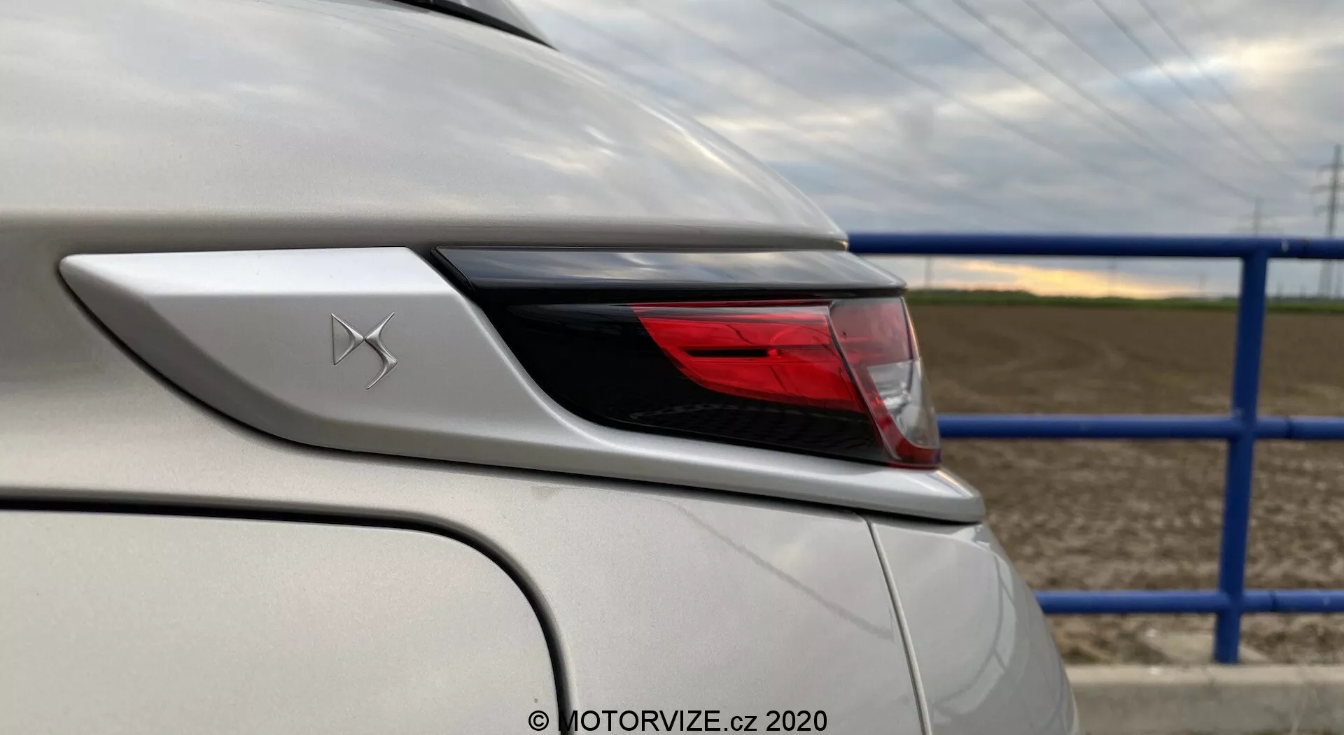 Detailný záber zadného rohu bieleho vozidla DS-3 2019 (DS-3 Crossback E-Tense), zameraný na dizajnové prvky a vlastnosti okolo zadného svetla. Uhol záberu zo zadného štvrtoblúkového uhla z ľavej strany zvýrazňuje zakrivenie zadného svetla a zadného blatníka, zobrazujúci detaily karosérie vozidla a pokročilé LED osvetlenie. Nad zadným svetlom je viditeľný odznak so štýlizovaným písmenom X, biely lak vozidla je v dokonalom stave a odráža lesklý povrch. Pozadie zahŕňa cestu s modrým kovovým plotom a poľnohospodárske pole.