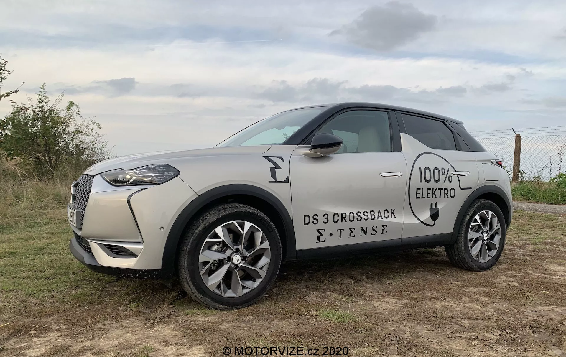 Bočný profil elektrického kompaktného SUV DS 3 Crossback E-Tense 2019 v pieskovom odtieni so kontrastnou čiernou strechou. Obrázok zachytáva prednú mriežku s chrómovým okrajom, LED svetlomety, odznaky '100% ELECTRO' a 'E-TENSE' na strane, viaclúčové zliatinové kolesá a zapustené kľučky dverí. Vozidlo je umiestnené vonku na trávnatom teréne pod zamračenou oblohou, čo naznačuje ranné alebo popoludňajšie osvetlenie.