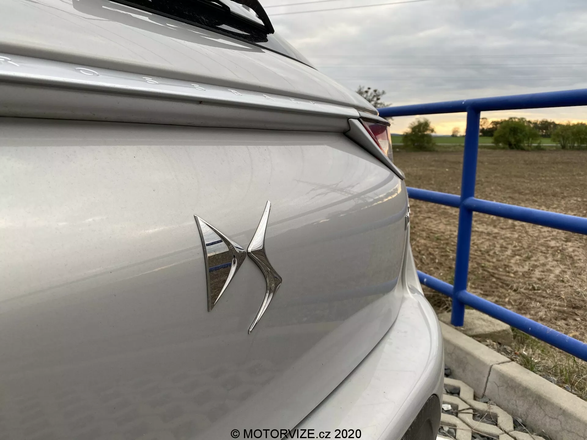 Detailný záber na zadnú časť luxusného sedanu alebo hatchbacku v svetlosivej alebo sivej farbe s výrazným emblémom značky na kufri a moderným LED zadným svetelným klastrom. Fotografia, vyhotovená pod miernym uhlom od zadnej časti smerom k pravej strane vozidla, ukazuje ostré horizontálne dizajnové línie a prímestské alebo vidiecke pozadie s modrými zábradlami a štruktúrami. Dizajnová estetika vozidla zdôrazňuje súčasnú technológiu a neutrálnu, tlmenú farebnú schému.