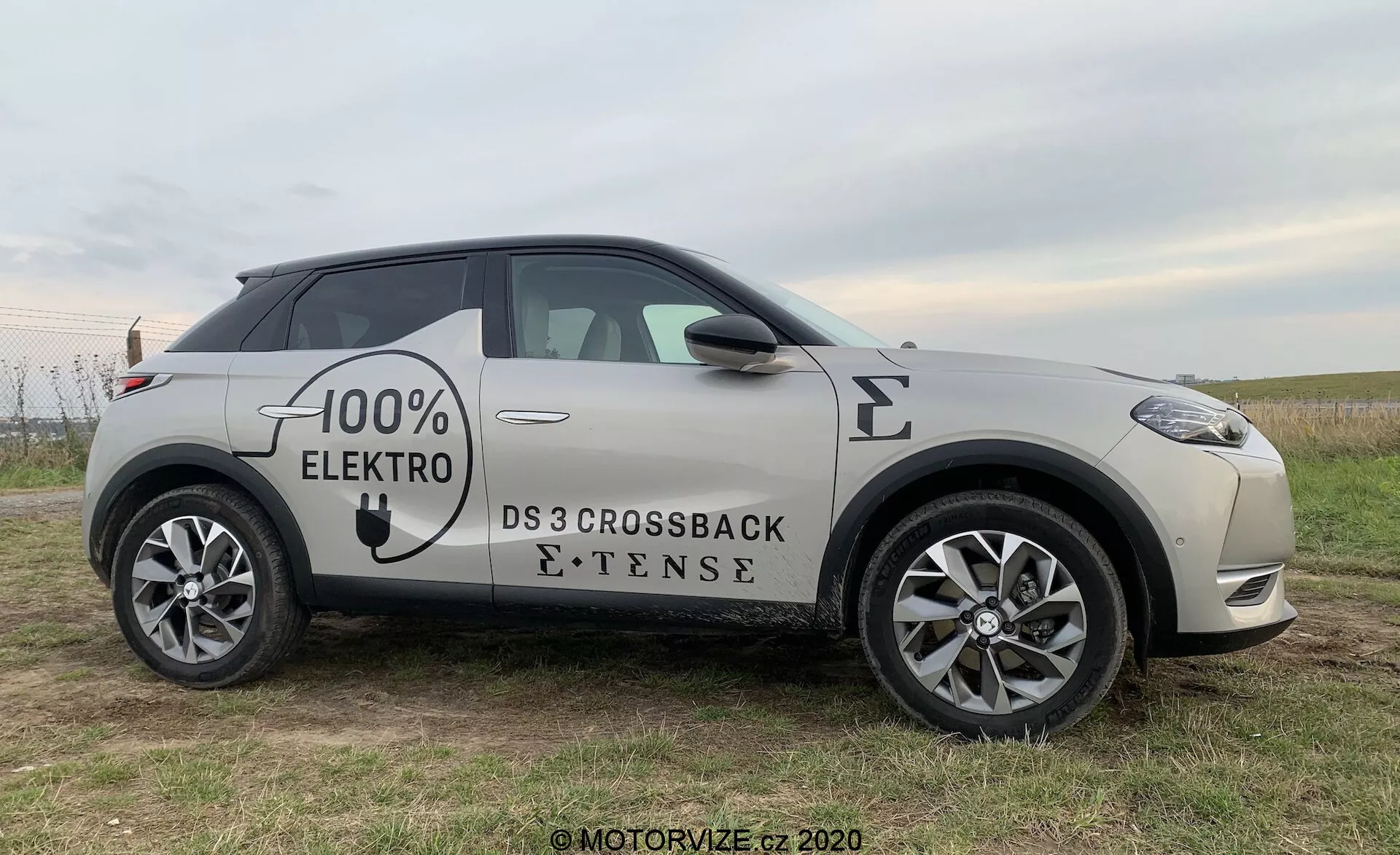 Bočný profil elektrického vozidla DS Automobiles DS 3 Crossback E-Tense z roku 2019, fotografovaný z mierneho predného uhla, ktorý zvýrazňuje predné koleso, dvere vodiča a zrkadlo, s čiastočnými pohľadmi na prednú mriežku a zadné koleso, umiestnený na trávnatom poli s jemným pozadím.
