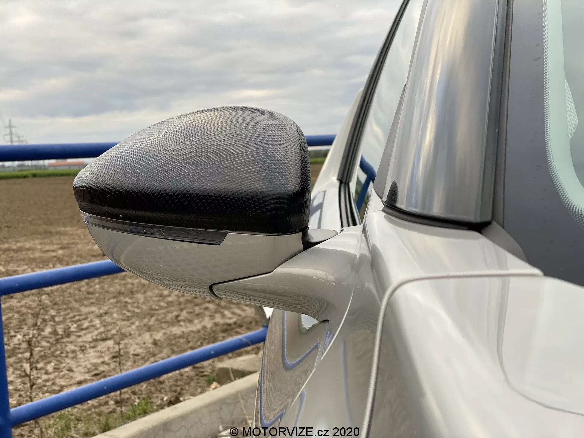 Blízky bočný pohľad na vonkajšie zrkadlo na dverách vodiča auta ds-automobiles ds-3 2019 (ds-3-crossback-e-tense). Zrkadlo je lakované tak, aby zodpovedalo farbe karosérie, okrem hornej časti krytu, ktorá má vzor z uhlíkových vlákien, čo naznačuje prémiový model. Pod zrkadlom je čiastočne viditeľný panel dverí s farbou karosérie a kontúrovou čiarou. Zrkadlo integruje moderné dizajnové prvky vrátane integrovanej LED smerovky. Obrázok sa zameriava na detaily dizajnu zrkadla s rozmazaným pozadím s otvoreným poľom a kovovou zábranou, čo naznačuje vidiecke prostredie.