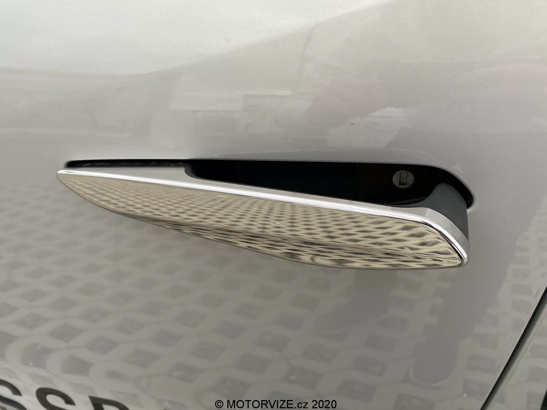Detailný pohľad na vetrací otvor vozidla DS Automobiles DS-3 2019 (DS-3 Crossback E-Tense) s elegantným chrómovým lemovaním a výrazným vlnitým vzorom vnútornej mriežky, zachytený z bočného uhla s jemným osvetlením pre zvýraznenie kriviek a dizajnových prvkov karosérie.