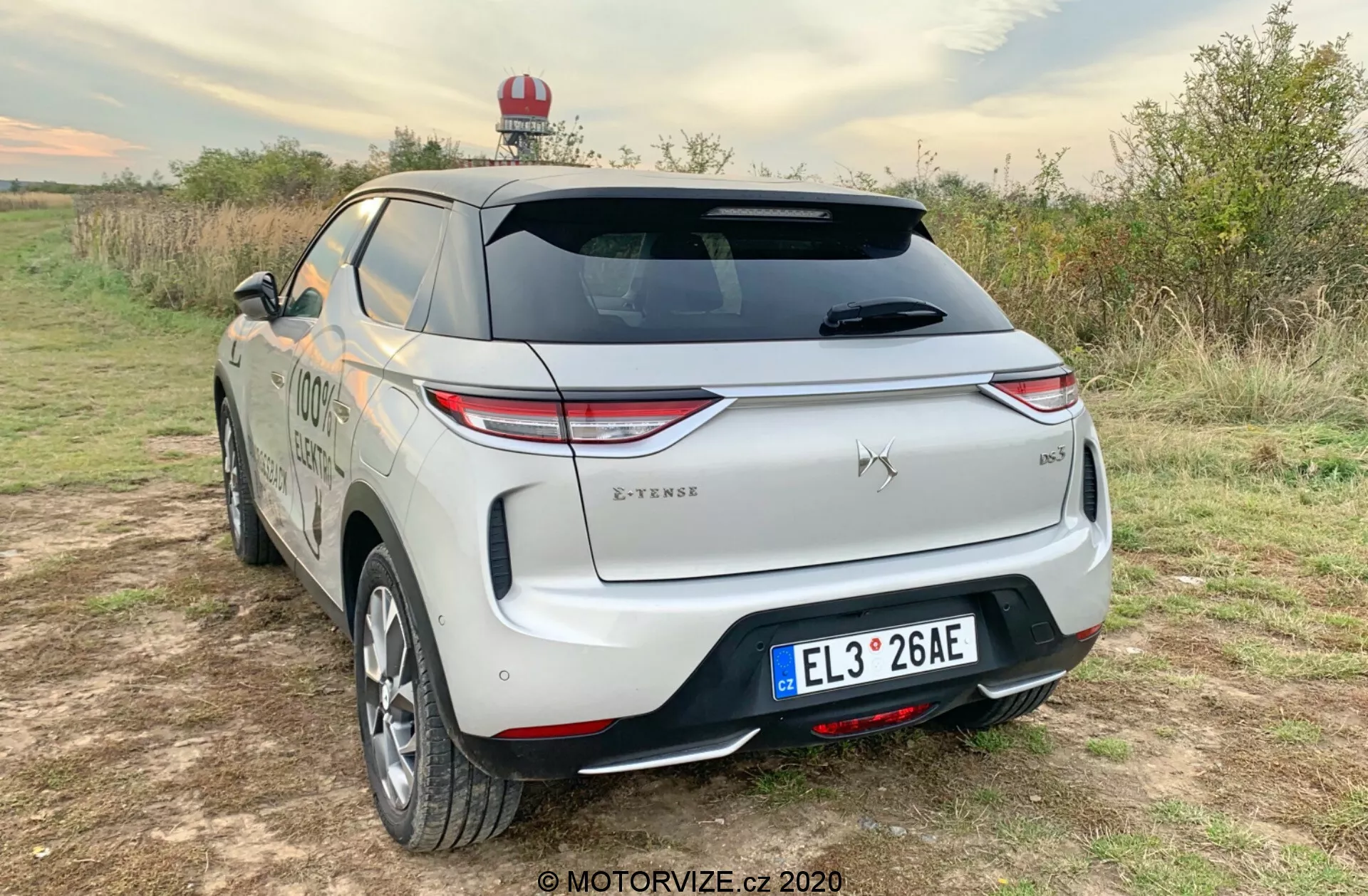 Trojštvrtinový zadný pohľad na SUV DS Automobiles DS-3 Crossback E-Tense 2019 zaparkované na neudržiavanej ploche v vidieckom prostredí, s LED zadnými svetlami, výrazným emblémom, zadným stieračom, výrazným zadným nárazníkom so senzormi, dvojfarebným povrchom, centrálnou poznávacou značkou, odznakom úrovne výbavy, strešným nosičom, možným núdzovým svetlom, hladkou aerodynamickou karosériou, zliatinovými kolesami, bočnými zrkadlami v karosárskom laku a nabíjacím krytom, ktorý naznačuje, že ide o elektrické alebo plug-in hybridné vozidlo.