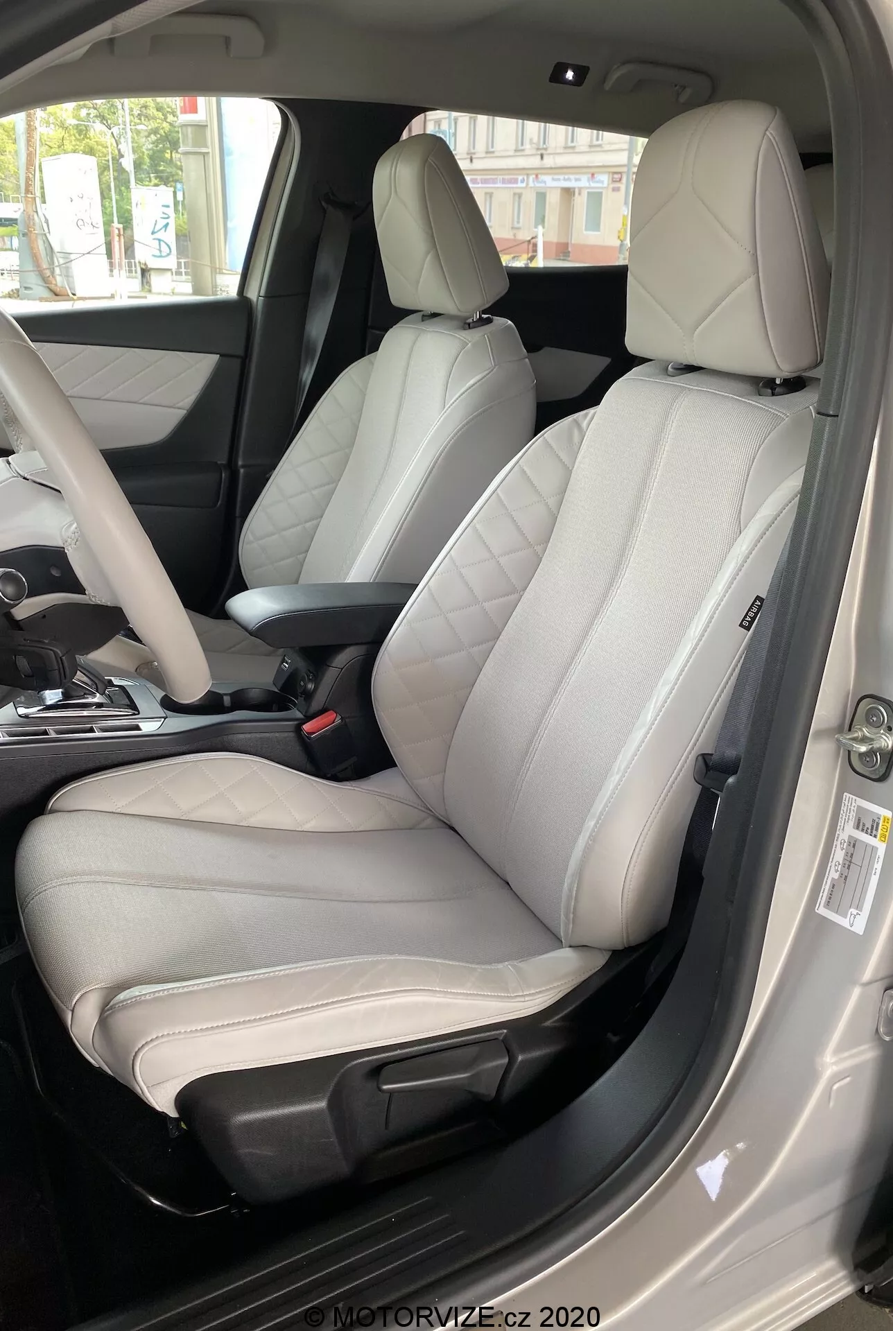 Interiér auta DS Automobiles DS 3 Crossback E-Tense 2019, zameraný na sedačku vodiča z vonkajších pohľadu na strane vodiča. Obrázok, fotografie z nízkeho uhla smerom nahor, ukazuje moderné dvojfarebné kožené sedadlo so svetlosivým stredom a tmavosivými alebo čiernymi okrajmi. Sedadlo je jednoznačne navrhnuté pre pohodlie a športový vzhľad, má výrazné bočné vystuženie, viditeľné šitie a elektronické nastavovacie ovládače. Otvorený vodičov dvere, viditeľný bezpečnostný pás, čiastočne viditeľná stredová konzola a panel dverí s rôznymi ovládacími gombíkmi naznačujú prvotriedne materiály a množstvo pokročilých funkcií. Denné svetlo zvýrazňuje jasnosť luxusných vnútorných detailov.