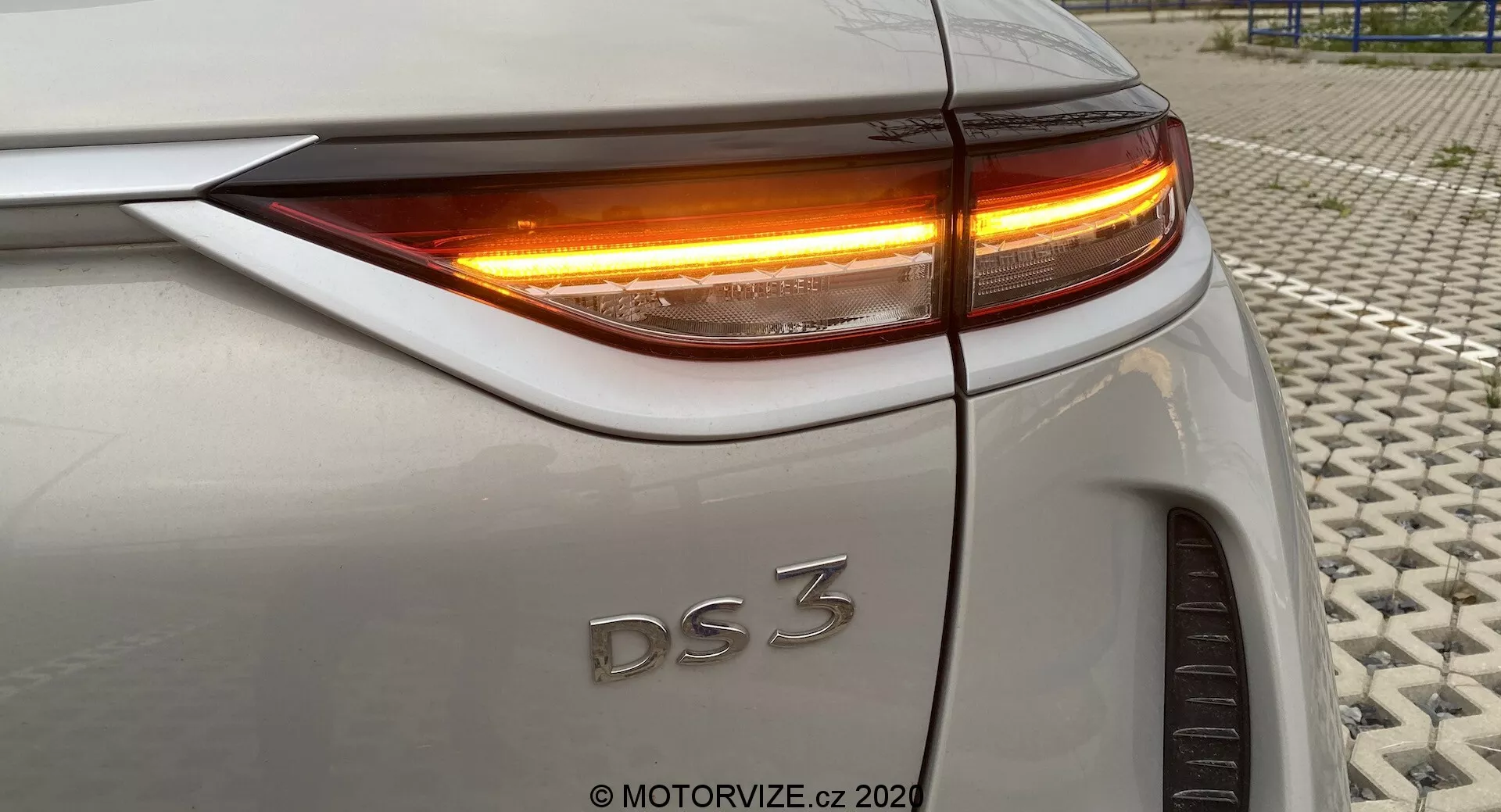 Detailný pohľad na zadnú časť DS 3 Crossback E-Tense (model 2019), ktorý ukazuje zadnú pravú štvrtinu z mierne vyvýšeného uhla. Obrázok zvýrazňuje LED zadné svetlo a smerovku s jasným oranžovým osvetlením. Viditeľné prvky zahŕňajú hornú hranu kufra, zakrivenie zadného blatníka, presné zarovnanie panelov karosérie a časť pravého zadného kolesa. Auto má striebornú alebo svetlosivú farbu. Fotografia bola odfotená na textúrovanom povrchu.