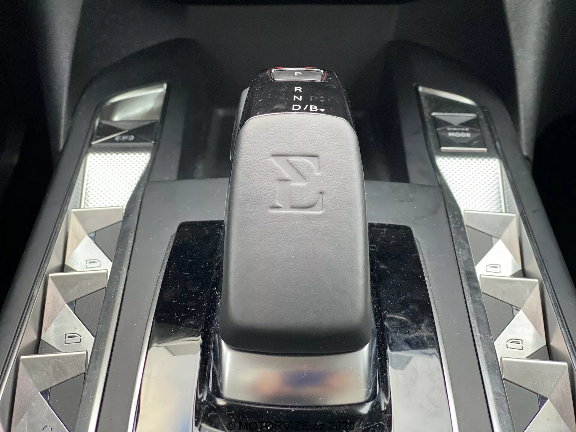 Detailný záber radiacej páky vozidla DS Automobiles DS-3 2019 (DS-3 Crossback E-Tense), zachytený z mierne vyvýšeného čelného uhlu. Radiaca páka, s elegantným čiernym koženým alebo koži podobným povrchom s indikátormi PRND, je centrom obrazu. Symetrické strieborné diamantové tlačidlá a dva kovové posuvníky po stranách naznačujú ďalšie funkcie vozidla. Vysoko lesklé a prémiové materiály vyžarujú moderný, luxusný interiér vozidla.