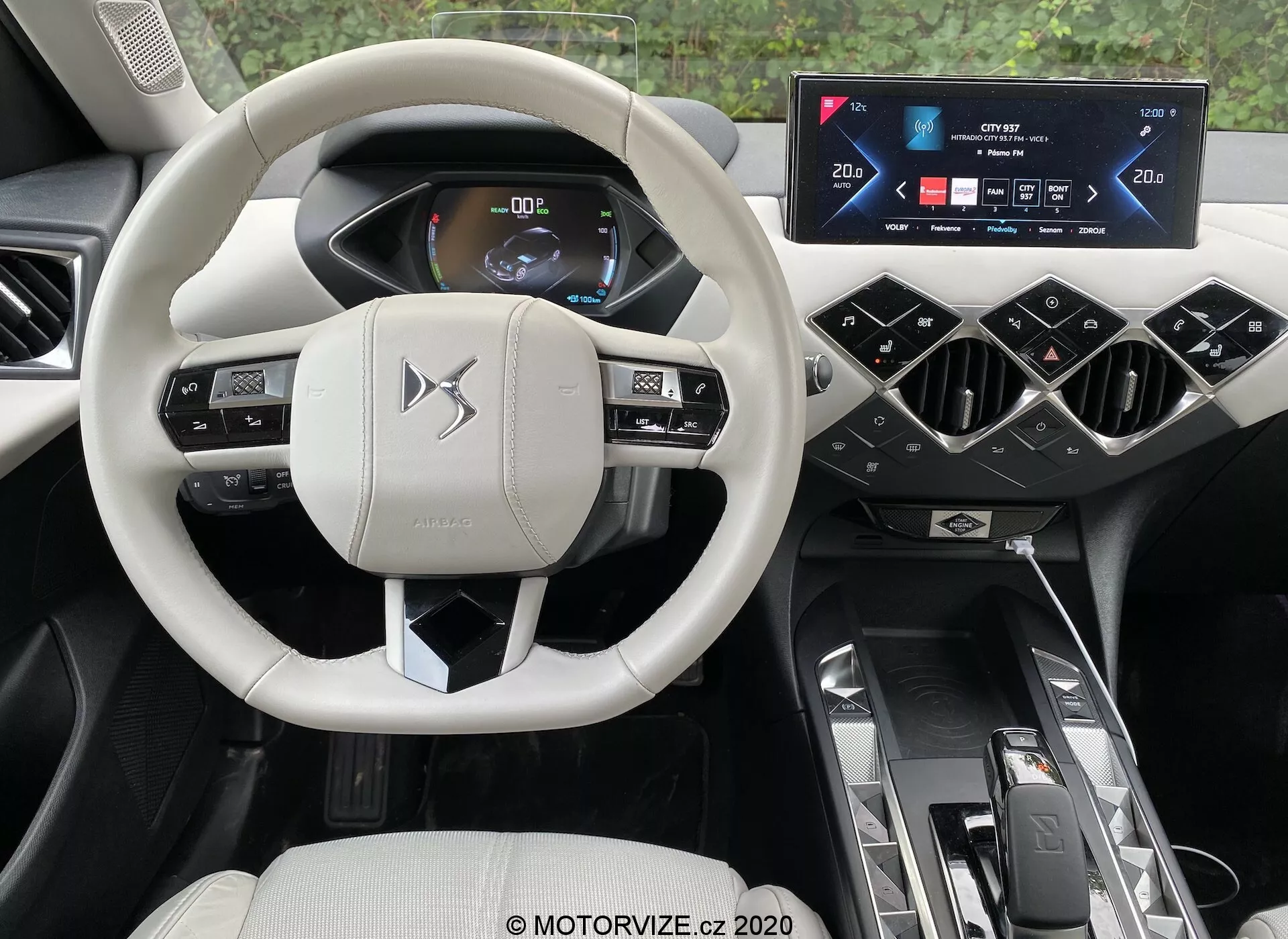 Pohľad do interiéru moderného automobilu z perspektívy vodiča, kde dominujú sofistikované prvky vozidla ds-automobiles ds-3 2019 (ds-3-crossback-e-tense). Uprostred je svetle zafarbený, multifunkčný volant s ovládacími tlačidlami a pádly, zasadený na pozadí digitálneho prístrojového panela zobrazujúceho informácie o vozidle s jasnou, čitateľnou grafikou. Nad stredovou konzolou je širokouhlý dotykový displej infotainmentu, po stranách ohraničený šikmými ventilačnými otvormi. Stredný tunel obsahuje automatický volič prevodovky, ovládanie režimov jazdy a dvojité držiaky na nápoje. Luxusný, prémiovej kvality interiér obsahuje svetlé a tmavé tóny, stelesňujúce komfort, pohodlie a pokročilé automobilové technológie.
