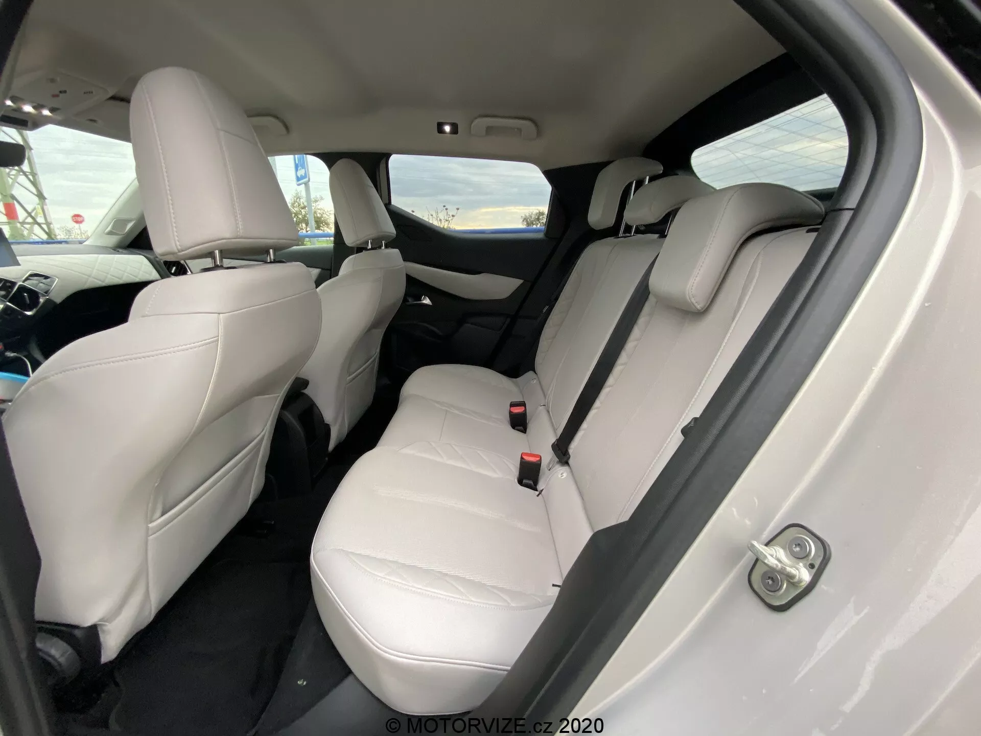 Interiér vozidla DS Automobiles DS 3 Crossback E-Tense 2019 z uhla, zameraného na zadný pasažiersky priestor. Svetlé kožené sedadlá, viditeľné opierky hlavy, bezpečnostné pásy a sklápateľná lakťová opierka na zadnom sedadle naznačujú luxusný a dobre udržiavaný interiér. Tmavé koberce kontrastujú so svetlým čalúnením, na dverách sú zreteľné ovládacie prvky elektrických okien. Uhol kamery, ktorý mierne smeruje nadol od zadných pravých dverí, zdôrazňuje oblasť sedadiel a priestor pre nohy.