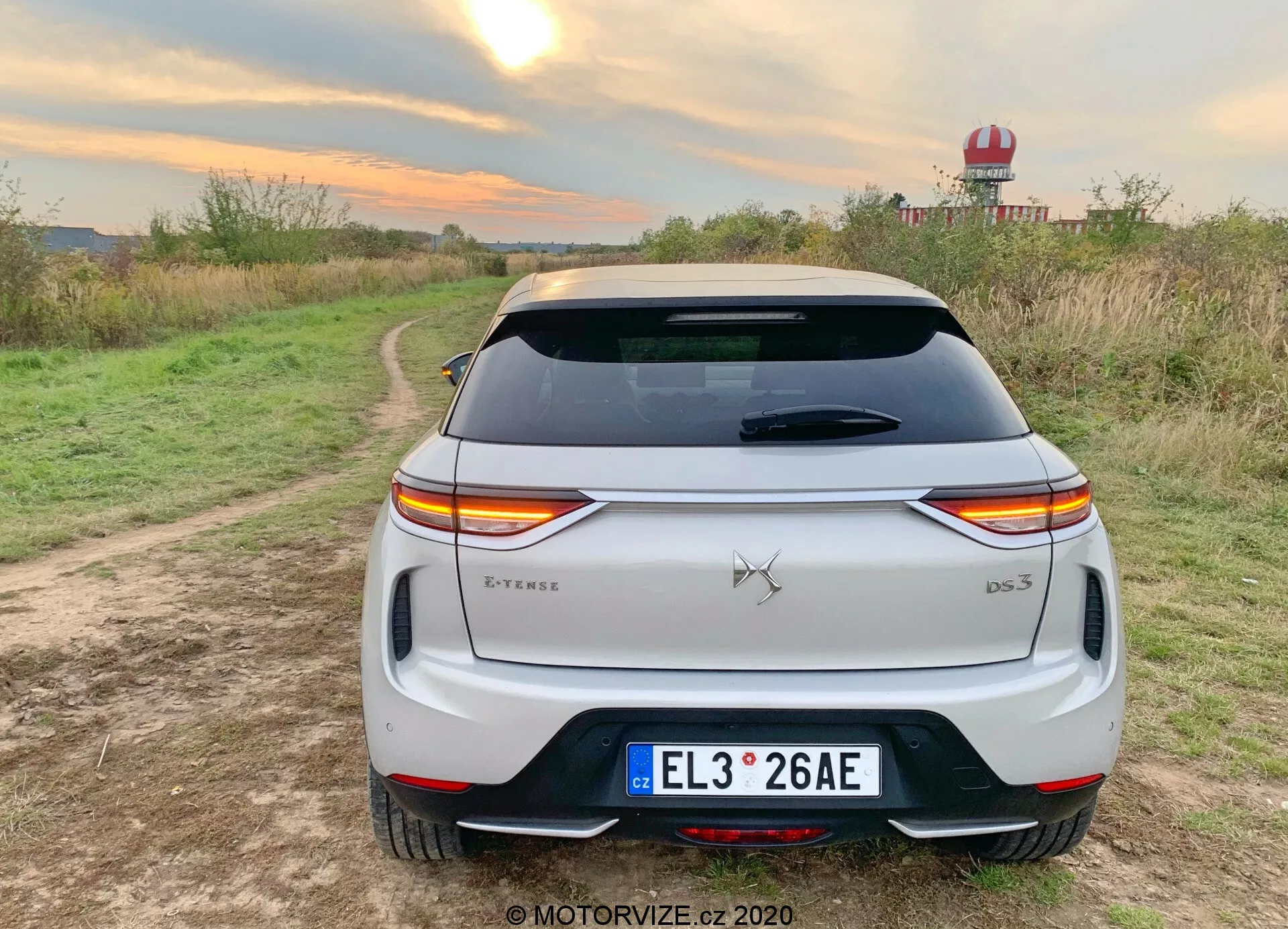 Zadný pohľad na biely 2019 DS Automobiles DS 3 Crossback E-Tense crossover SUV zaparkovaný na štrkovej ceste s vegetáciou po oboch stranách v vidieckom prostredí. Charakteristické horizontálne orientované LED zadné svetlá sa tiahnu naprieč zadnou časťou vozidla, prerušované emblémom značky. Nápis modelu 'DS 3' sa nachádza v pravom dolnom rohu kufra, s odznakom 'E-Tense' na ľavej dolnej strane, čo naznačuje elektrický alebo hybridný pohon. Vlastnosti zahŕňajú európsku poznávaciu značku, zadné sklo s odmrazovacími linkami, strešný spojler, zadný nárazník s integrovanými reflektormi a čierny lem, a dvojité výfukové koncovky, čo naznačuje športovejší dizajn. Prostredie naznačuje skúšobnú jazdu alebo životný štýlový fotografický záber zdôrazňujúci vonkajšie použitie, s teplými svetelnými podmienkami zdôrazňujúcimi bielu farbu vozidla.
