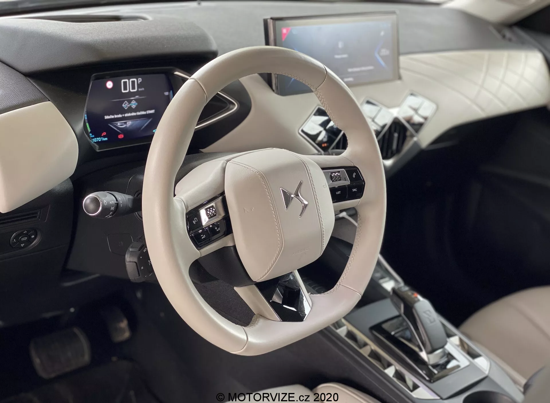 Záber interiéru vozidla DS-Automobiles DS-3 2019 (DS-3 Crossback E-Tense) zameraný na svetlý volant s emblémom Mitsubishi a stredovú konzolu. Fotografia z perspektívy zadných sedadiel ukazuje ovládacie prvky pre audio, hlas a tempomat. Digitálny prístrojový panel zobrazujúci nulovú rýchlosť a páka radenia v polohe 'P', naznačujúce, že auto stojí. Viditeľný centrálny infotainment systém s dotykovou obrazovkou, situovaný v dvojfarebnom interiéri s kovovými akcentmi, naznačujúcimi moderné digitálne displeje a pokročilé automobilové funkcie.