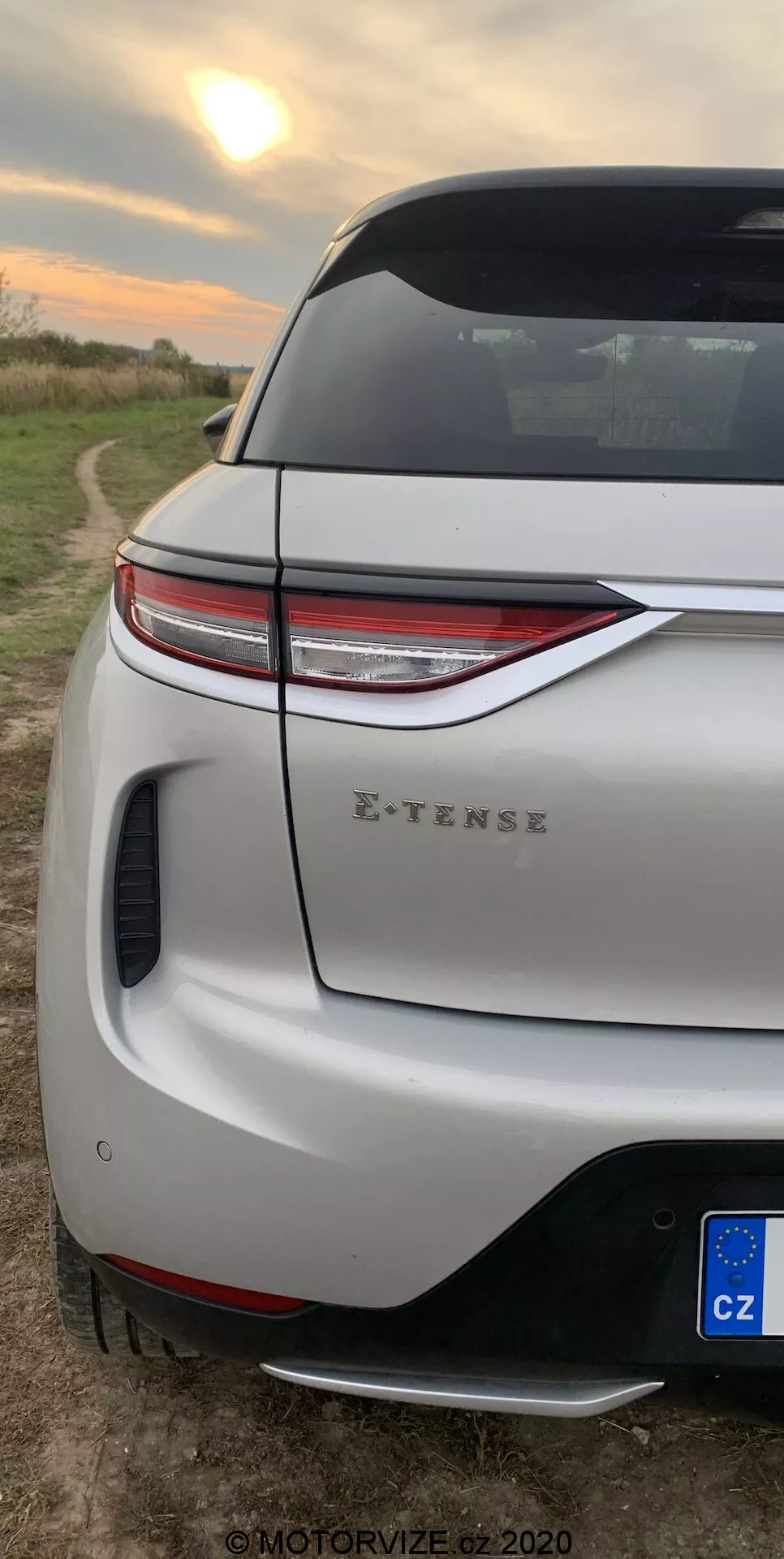 Vertikálny detailný záber zadnej rohovej časti vozidla DS-3 2019 (DS-3 Crossback E-TENSE). Zameriava sa na moderné LED zadné svetlá, športový zadný nárazník s integrovaným difúzorom a parkovacím senzorom, veku kufra s aerodynamickým spojlerom a čiastočný pohľad na zadné koleso a blatník. Nachádza sa na vidieckom pozadí s otvoreným poľom pri východe alebo západe slnka, čo naznačuje moderný dizajn vozidla a jeho elektrické schopnosti.