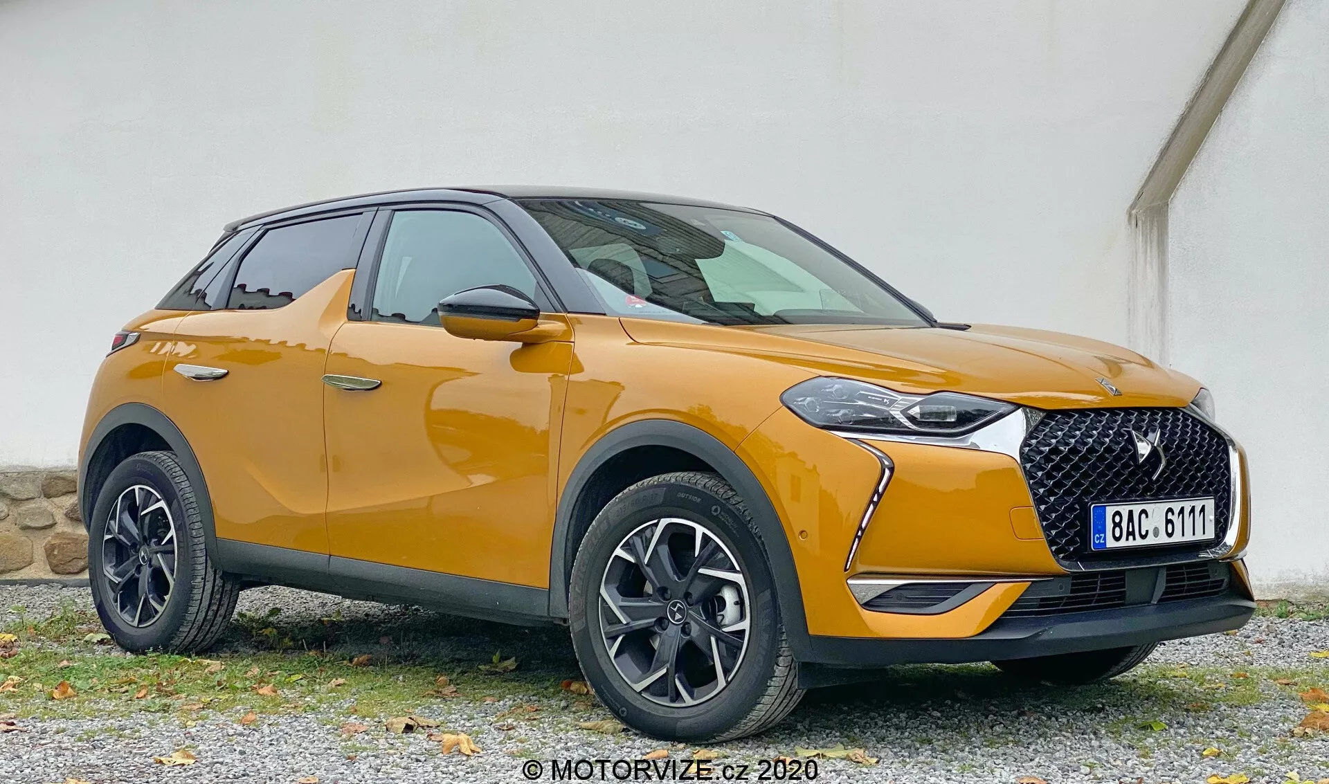 Moderné SUV v trojštvrťovom prednom pohľade, zobrazujúce DS Automobiles DS 3 2019 (DS 3 Crossback) s detailmi ľavého predného rohu vrátane reflektora, mriežky, kapoty, predného nárazníka, kolesa, predného blatníka a dverí vodiča, umiestnené na štrkovom povrchu so zoslabenou farebnou stenou v pozadí.
