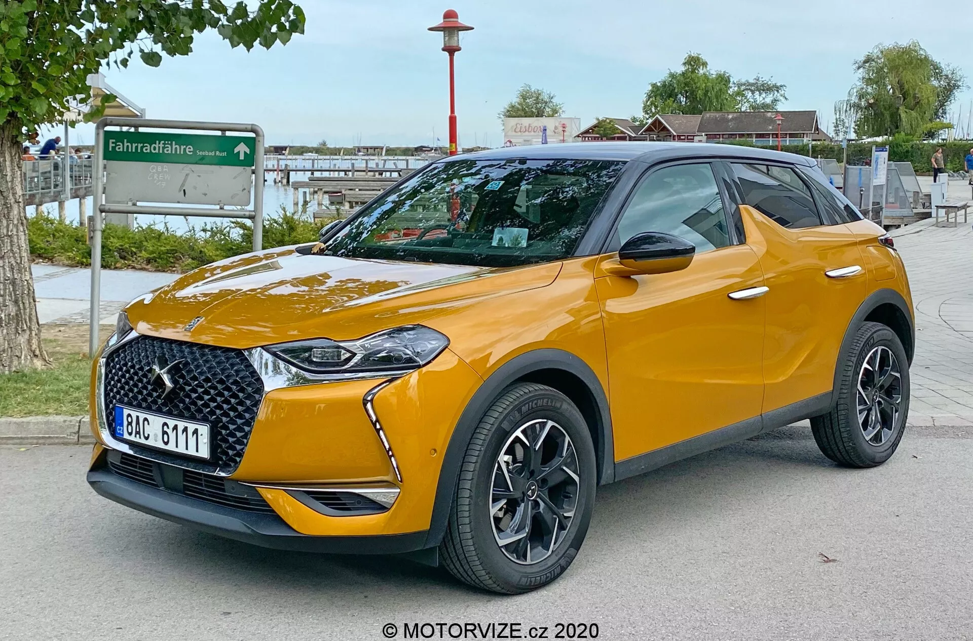 Obrázok zobrazujúci moderné SUV DS Automobiles DS 3 Crossback 2019 vo výhľade z troch štvrtín zpredu, zdôrazňujúci štýlovú prednú mriežku, integrované LED svetlomety, predný nárazník, kapotu a bočný profil vodiča. Auto je zaparkované vonku na spevnenej ploche s mestským pozadím, ktoré zahŕňa stanicu na zdieľanie bicyklov. Fotografiu nasnímanú z nízkeho uhla vo výške svetlometov zvýrazňuje svetlú výšku vozidla. Viditeľné vlastnosti zahŕňajú viaclúčové zliatinové kolesá, integrované smerovky v zrkadlách, nízkoprofilové pneumatiky, kontrastnú farbu strechy so strešnými nosičmi, metalický lak, odznaky špecifické pre trim a tónované zadné okná. ŠPZ je úmyselne rozmazaná pre ochranu súkromia.