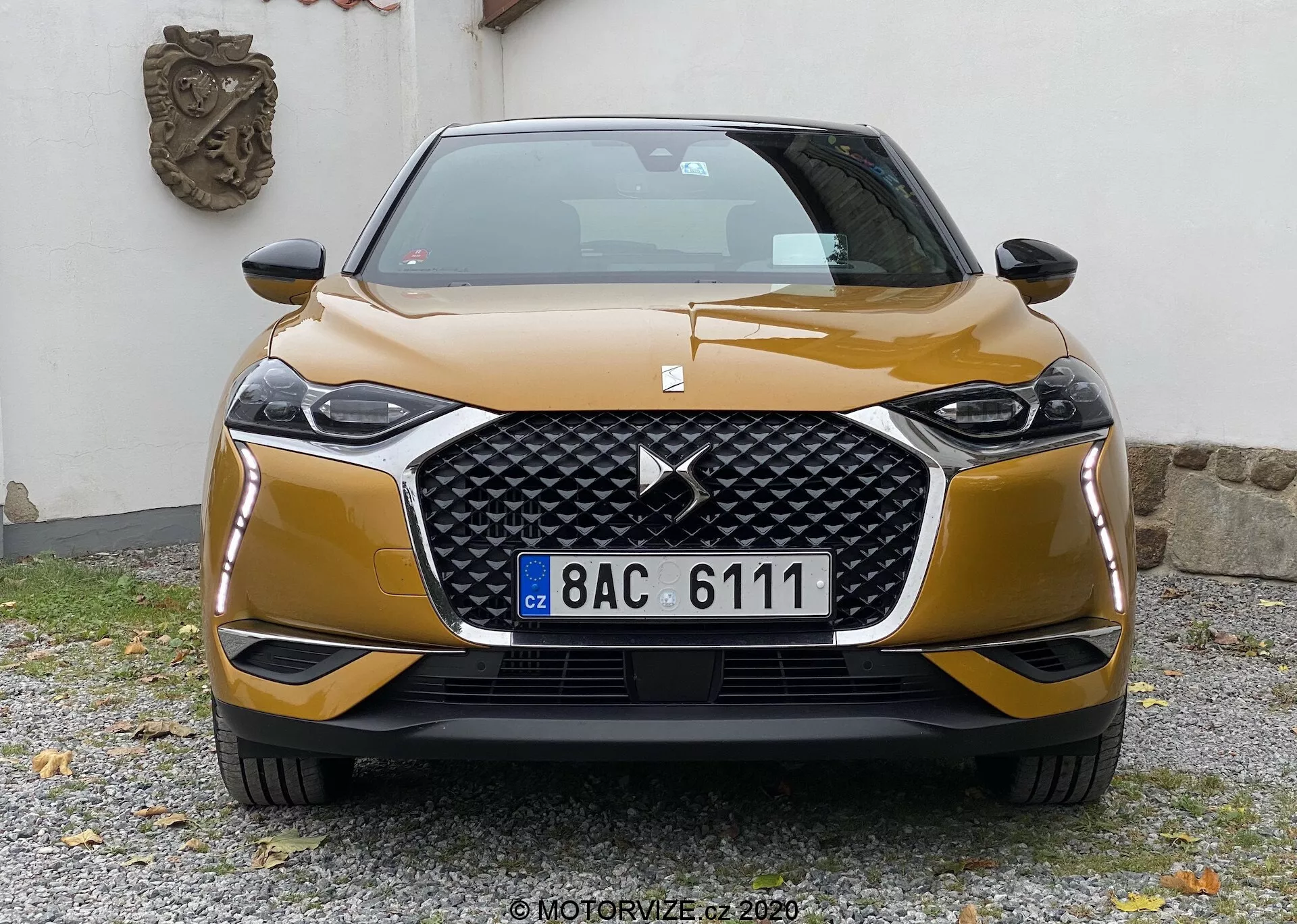 Pohľad spredu na vozidlo DS-3 Crossback 2019, so zreteľným šesťuholníkovým grilom s veľkým logom v strede, obklopený elegantnými uhlovými svetlometmi pravdepodobne s LED alebo HID technológiou. Športový predný nárazník s kontrastným obložením a tvarovanými líniami na kapote pridávajú dynamiku estetiky. Mierne posunuté auto je fotografované vo výške grilu, zachytávajúce moderný dizajn a technológie.