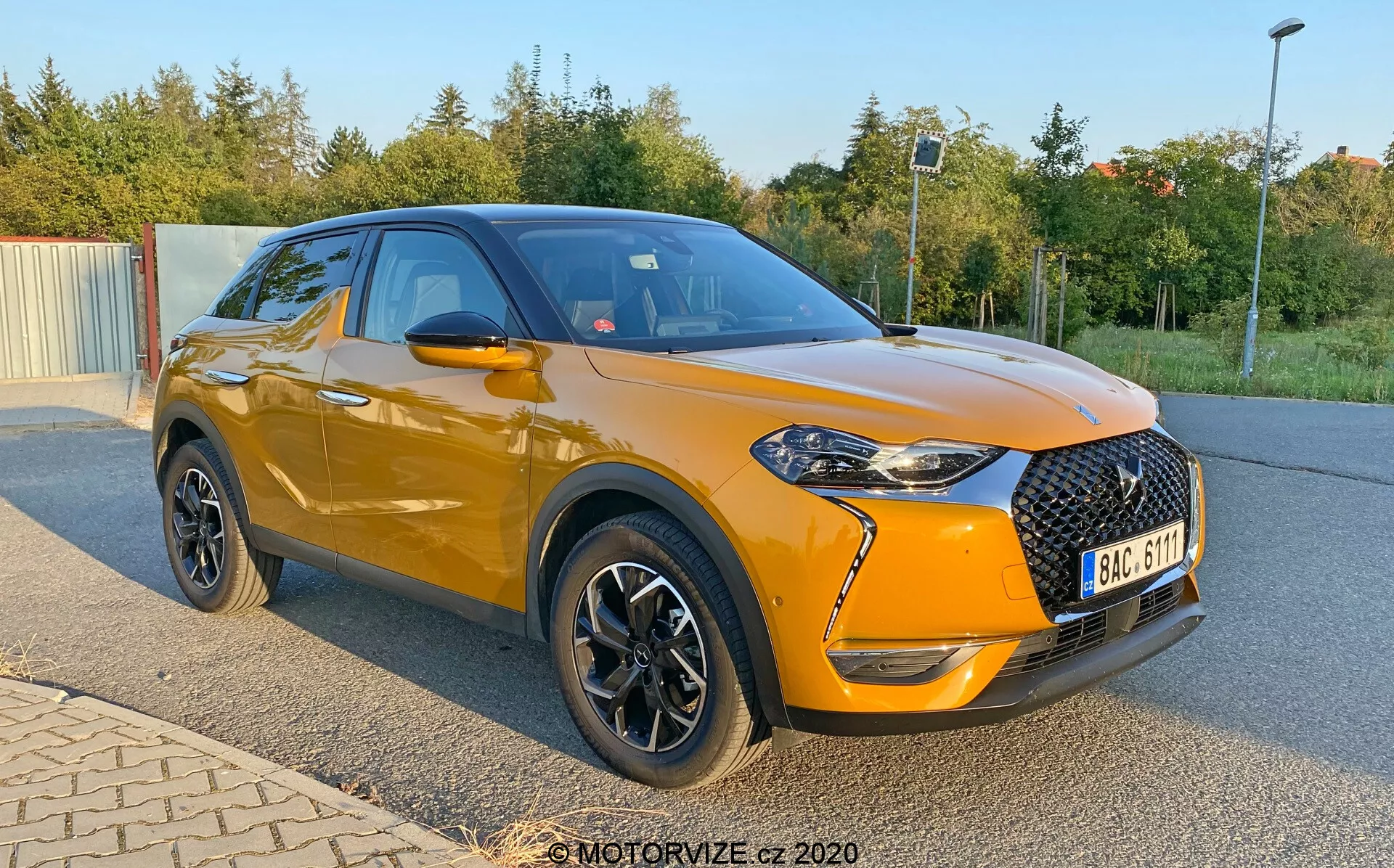 Moderné, kompaktné SUV DS Automobiles DS-3 Crossback z roku 2019, ukázané zaparkované pod uhlom s dôrazom na predný a bočný profil. Obrázok, zachytený z nižšej perspektívy blízko nábojov kolies, zvýrazňuje jasne oranžové kovové telo vozidla a čiernu strechu. Predná časť obsahuje veľkú čiernu mriežku s diamantovým vzorom, ostré svetlomety s LED akcentmi a hmlové svetlá v čiernych rámoch. Auto má čierne spätné zrkadlá, chrómové okenné lišty a čierne kolesá so striebornými akcentmi. Nápadný je mierne vyšší postoj, športové línie tela a klesajúci záhyb cez kľučky dverí.