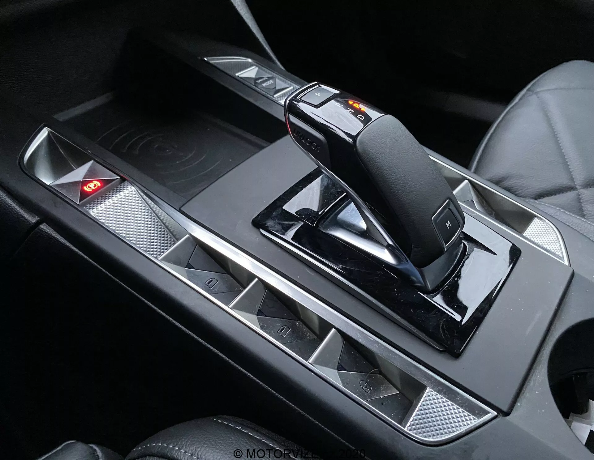 Detailný záber interiéru DS Automobiles DS-3 Crossback 2019, zameraný na stredovú konzolu s modernou, ergonomickou radiacou pákou s prepínačom manuálneho režimu a uvoľňovacím mechanizmom. Konzola má kovové akcenty, ktoré kontrastujú s čiernymi povrchmi, držiaky na nápoje s matným povrchom a ovládací panel s červeným výstražným svetlom.