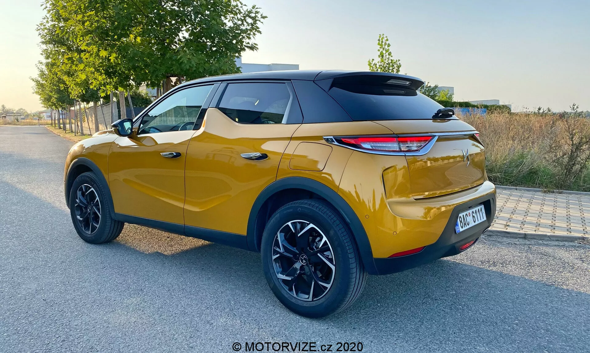 Zadný trojštvrtinový pohľad na moderné SUV crossover za denného svetla, so žiarivou metalickou zlatou farbou a športovou čiernou strechou, modernými LED zadnými svetlami, veľkými čiernymi zliatinovými kolesami, vysokou svetlosťou, viditeľnými zadnými parkovacími senzormi a dvojfarebným dizajnom, typickým pre model DS Automobiles DS 3 Crossback z roku 2019.