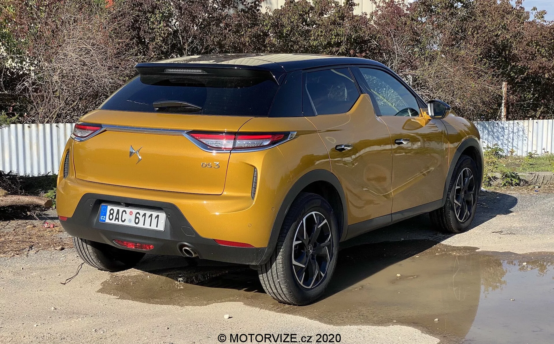 Pohľad zozadu na kovový zlatý DS-3 Crossback SUV zaparkovaný vonku na slnečný deň. Obrázok zachytáva vozidlo z mierne pravého uhla a zvýrazňuje pravú zadnú stranu a oblasť kufra. Kľúčové vlastnosti zahŕňajú moderné hranaté zadné svetlomety, šikmé zadné sklo s integrovaným strešným spojlerom, výrazný nárazník s parkovacími senzormi, čierne viacramenné zliatinové disky, vysokú svetlú výšku a zatmavené zadné okná.