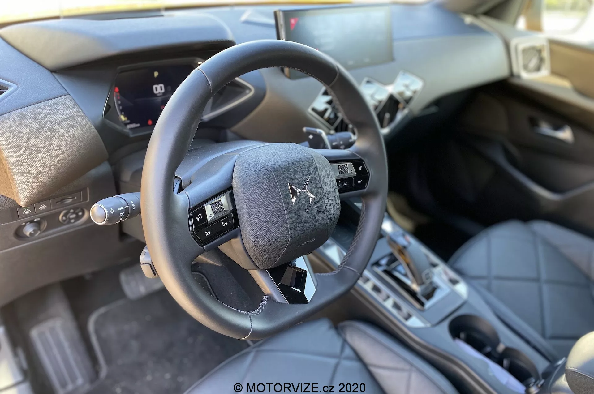 Pohľad do interiéru DS Automobiles DS 3 Crossback 2019 z perspektívy predného sedadla spolujazdca, zvýrazňujúci trojramenný volant s multifunkčnými tlačidlami, čiastočný digitálny prístrojový panel zobrazujúci rýchlosť auta a merače, stredovú konzolu s automatickou prevodovkou, prémiové materiály ako kožené povrchy a kovové akcenty, tradičný slot pre zapínanie zapaľovania, pedále a dobre osvetlené prirodzeným denným svetlom.
