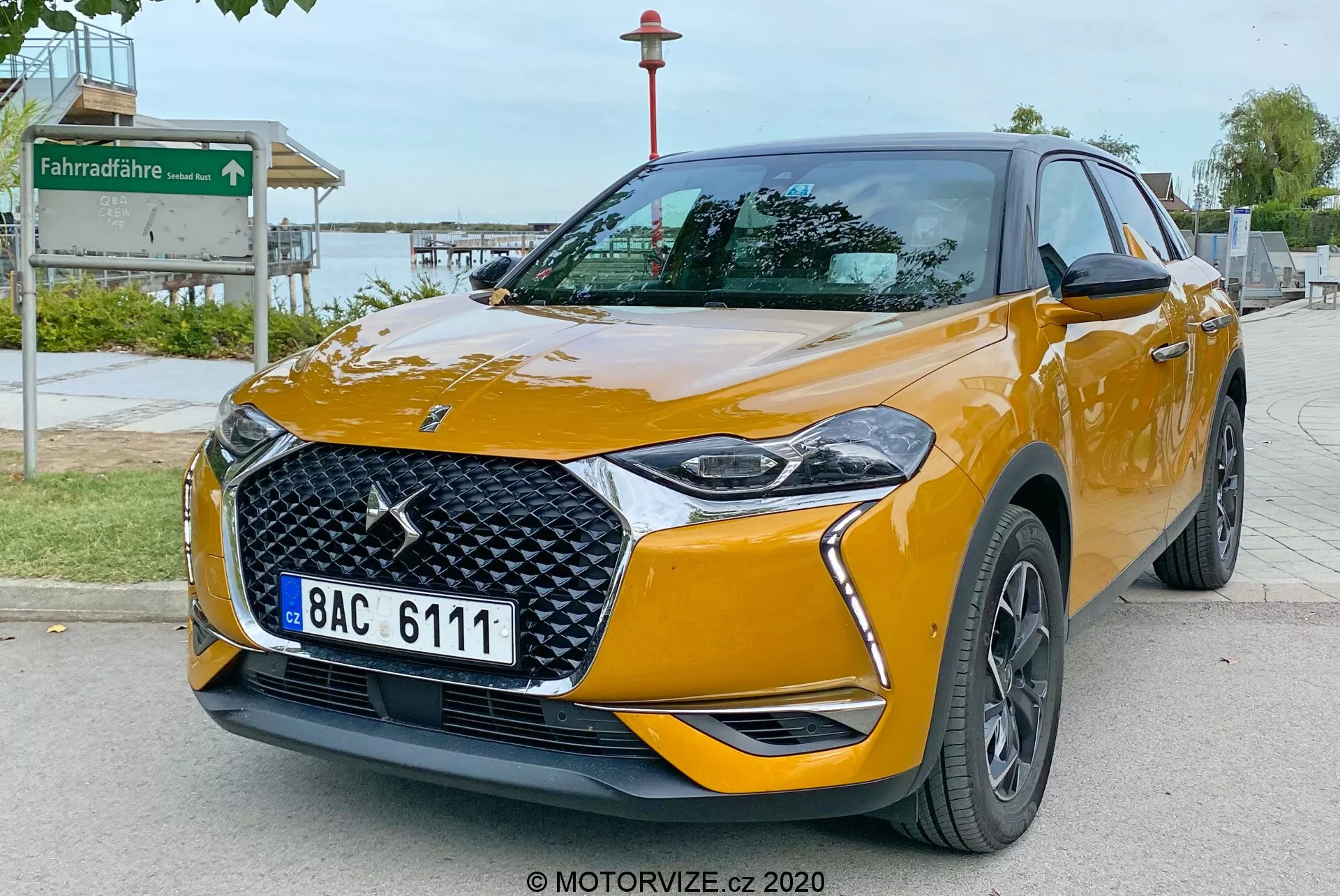 Obrázok zobrazuje DS Automobiles DS 3 Crossback z roku 2019, moderný kompaktný crossover SUV, zaparkovaný na dláždenom chodníku. Zhotovený z nízkeho trojštvrťového uhla vpredu, obraz zdôrazňuje prednú časť vozidla a jeho pravú stranu. Kľúčové prvky zahŕňajú výrazné logo značky na mriežke s diamantovým vzorom, elegantné LED svetlomety so štýlovými dennými svetlami, integrované hmlové svetlá v prednom nárazníku, zrkadlá vo farbe karosérie s ukazovateľmi smeru a tmavé viacramenné zliatinové kolesá. Auto je natreté výraznou leskle oranžovou farbou s výraznými líniami karosérie, ktoré zlepšujú jeho aerodynamický vzhľad, a odznaky, ktoré môžu naznačovať úrovne výbavy. Okolie naznačuje verejnú oblasť v blízkosti parku, ktorú naznačuje znak cyklistickej cestičky v pozadí.