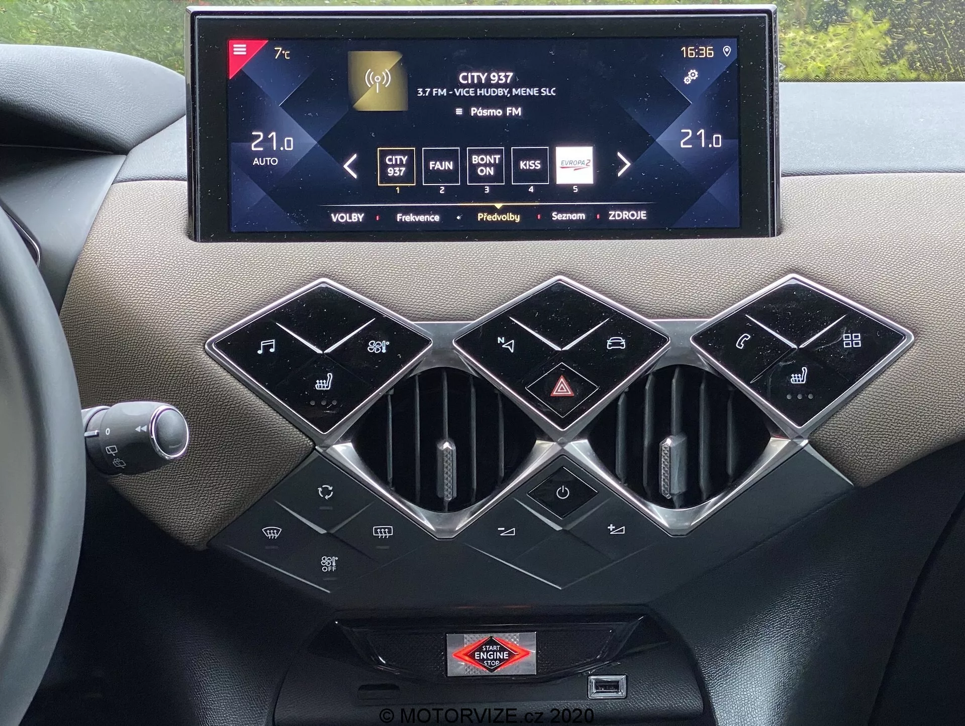 Interiérový pohľad na stredovú konzolu ds-automobiles ds-3 2019 s veľkým dotykovým infotainment systémom zobrazujúcim ovládanie klimatizácie, informácie o rádiu a nastavení, s tromi šesťhrannými vetracími otvormi pod ním s lesklým strieborným lemovaním. Pod nimi sa nachádza rad čiernych lesklých tlačidiel na ovládanie klimatizácie, vrátane tlačidla SYNC pre viaczónovú klimatizáciu, automatickej klimatizácie a značkového znaku umiestneného pod dotykovou obrazovkou, zvýrazňujúce prémiové hmatové materiály a modernú automobilovú technológiu