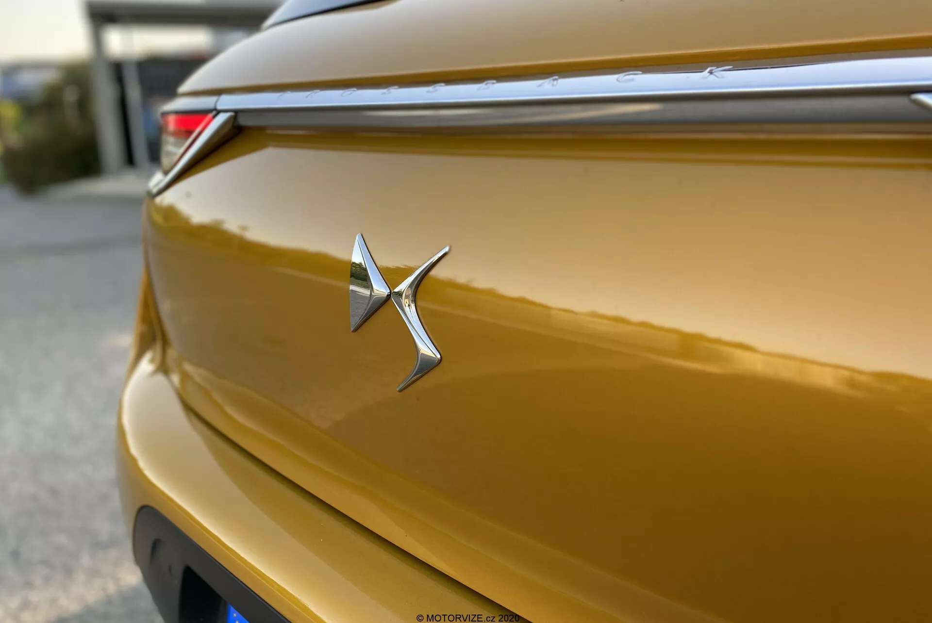 Detailný záber na kovový zlatý znak DS Automobiles DS-3 Crossback 2019. Chrómový znak je ostrý a centrálne umiestnený, pripomínajúci hviezdu s predĺženým pravým ramenom. Lesklá karoséria odráža svetlo na svojich krivkách, ukazujúc bohatý zlatý odtieň. Plytká hĺbka ostrosti zvýrazňuje znak, s horizontálnou čiarou nad ním prispievajúcou k aerodynamickému dizajnu. V pravom hornom rohu je čiastočne viditeľné zadné LED svetlo. Zadné veko má jemné zakrivenie a efekt bokeh v pozadí izoluje znak.