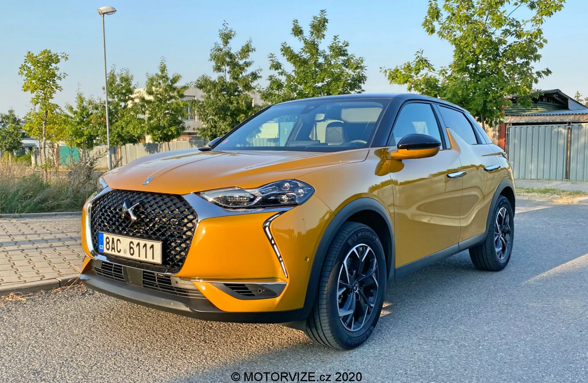 Predný trojštvrťový pohľad na DS Automobiles DS 3 Crossback z roku 2019, ktorý ukazuje výraznú veľkú prednú mriežku so sieťovým vzorom a výrazným znakom, elegantné LED svetlomety, metalickú oranžovú farbu, kontrastné čierne zliatinové kolesá, integrované bočné zrkadlá a stúpajúcu líniu okien končiacu aerodynamickým spojlerom. Vozidlo je zaparkované so zeleným lístím a štruktúrami v rozmazanom pozadí, čo zdôrazňuje dizajn a športové vlastnosti auta.