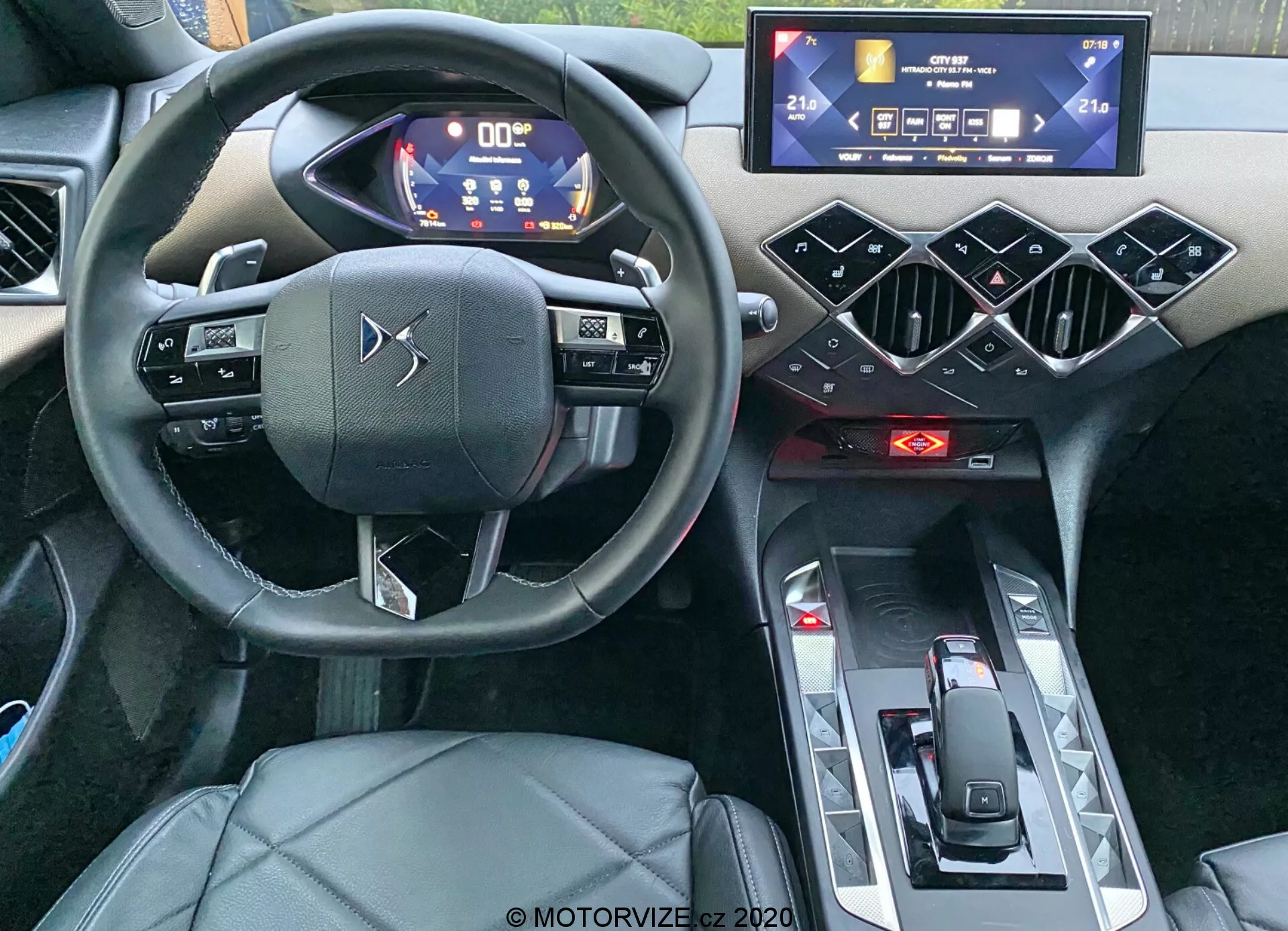 Pohľad do interiéru DS Automobiles DS-3 Crossback 2019 zameraný na kokpit vodiča z úrovne očí. Záber urobený z oblasti zadného pasažiera zvýrazňuje trojramenný volant s kapacitnými dotykovými ovládačmi, digitálny prístrojový panel s modrým a bielym osvetlením, horizontálne orientovaný dotykový infotainment systém, geometrické šesťuholníkové dizajnové prvky na stredovej konzole, červené indikátory klimatizácie, minimalistický volič prevodových stupňov a sedadlo vodiča potiahnuté kožou alebo syntetickou kožou. Fotografia zdôrazňuje moderné, vysoko technické prvky a luxusný dizajn auta.