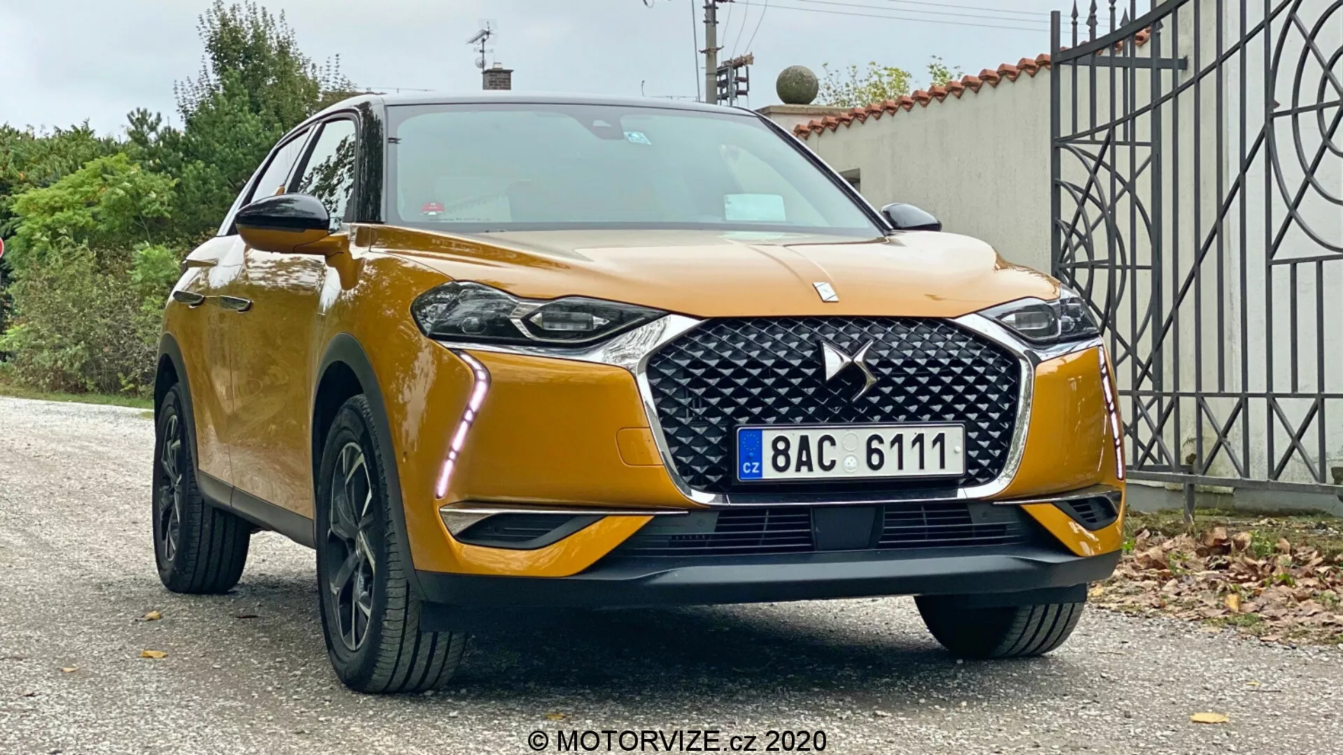 Predný trojštvrťový pohľad na SUV DS Automobiles DS 3 Crossback z roku 2019, ktorý ukazuje jeho výšku a robustné držanie tela na predmestskom pozadí. Viditeľné sú kovová zlatá farba, výrazná mriežka chladiča s diamantovým vzorom a logom, elegantné LED svetlomety, hmlové svetlá, mohutný predný nárazník so širokým nasávacím otvorom, čierne viacramenné zliatinové disky a bočné zrkadlá s integrovanými smerovkami.