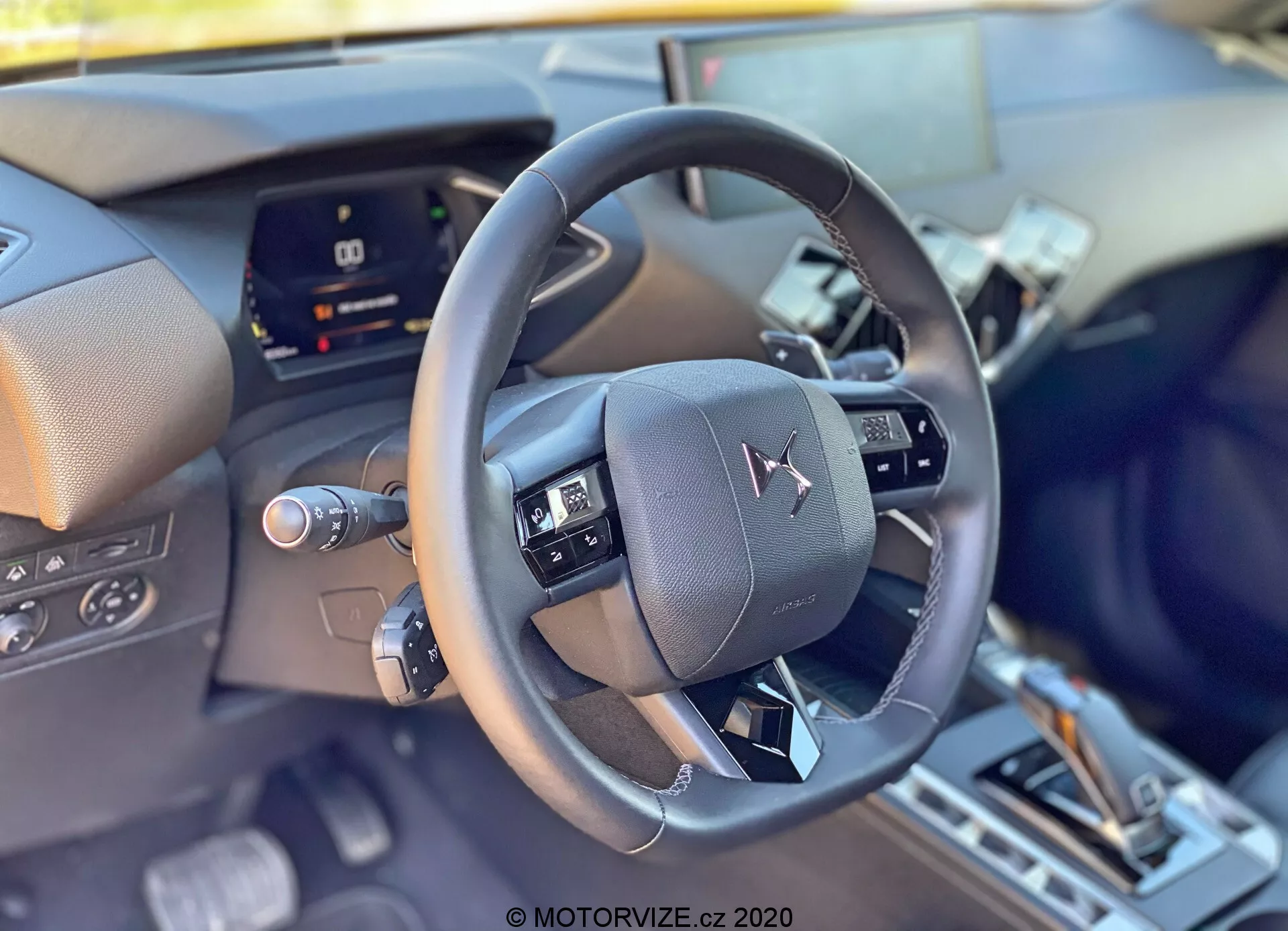Detailný pohľad na interiér DS Automobiles DS-3 2019 (DS-3 Crossback) z pohľadu vodiča, zameraný na moderný kožený volant v strede obrazu s multifunkčnými ovládacími prvkami. Za volantom sa nachádza digitálny prístrojový panel, čiastočne zakrytý, naznačujúci pokročilú technológiu vozidla. V dolnej časti je náznak stredovej konzoly s radiacou pákou a možnými voliči jazdných režimov, čo odráža sofistikované interiérové a komfortné prvky vozidla.