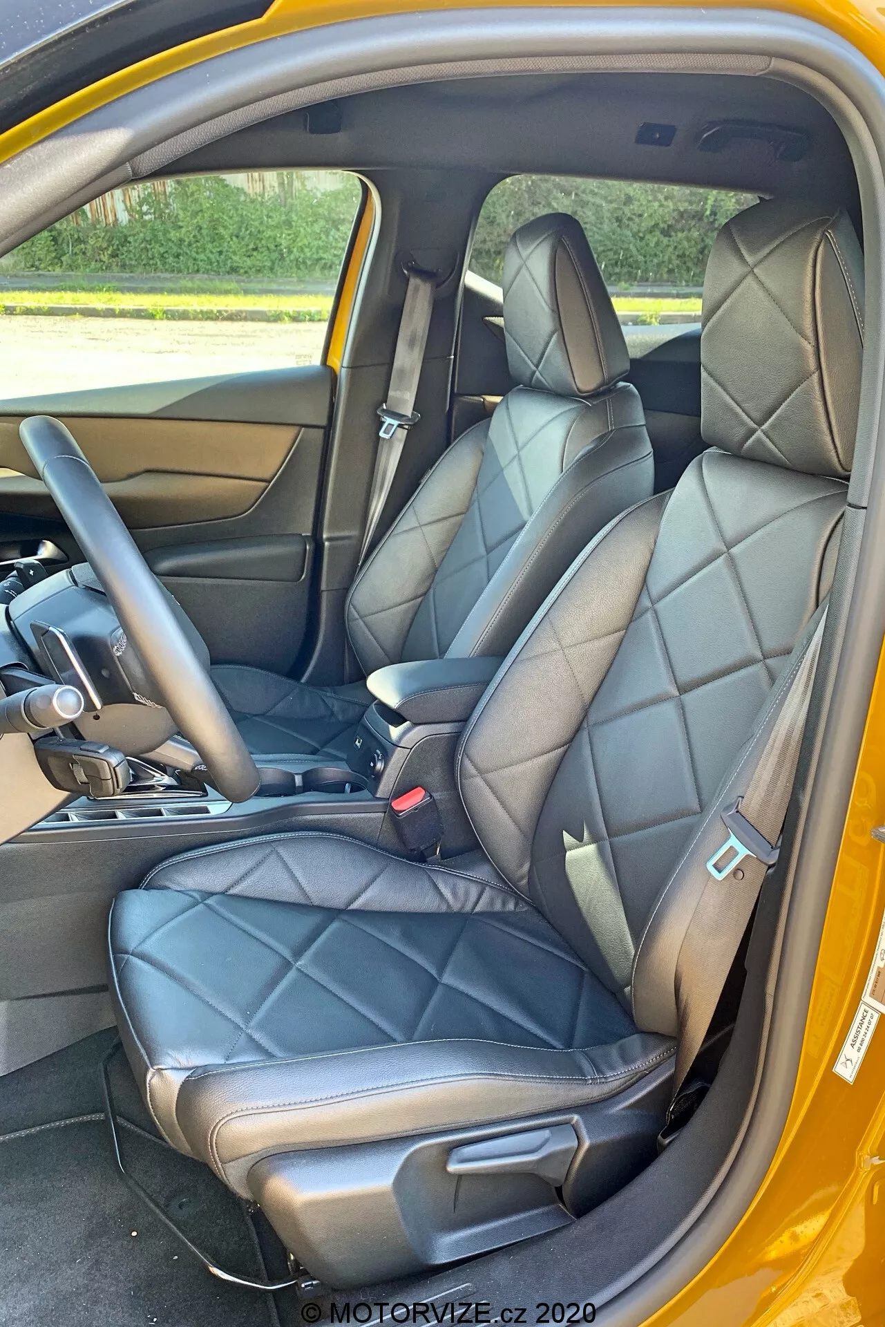 Pohľad na interiér DS Automobiles DS-3 Crossback 2019 zameraný na predné sedadlá z pohľadu z bočných dverí spolujazdca. Obrázok zvýrazňuje čierne kožené prešívané sedadlá so zabudovanými opierkami hlavy a bočnými oporami, čo naznačuje luxusný alebo športový dizajn. Viditeľná je časť stredovej konzoly so radiacou pákou naznačujúcou automatickú prevodovku a dolná časť palubnej dosky. Vnútorný panel dverí zodpovedá koži sedadiel, s kovovou kľučkou a ovládacími tlačidlami. Viditeľné sú aj čierne bezpečnostné pásy s mechanizmom na nastavenie výšky, kobercová podlaha a kontrastná vonkajšia farebná schéma, pravdepodobne žltá.