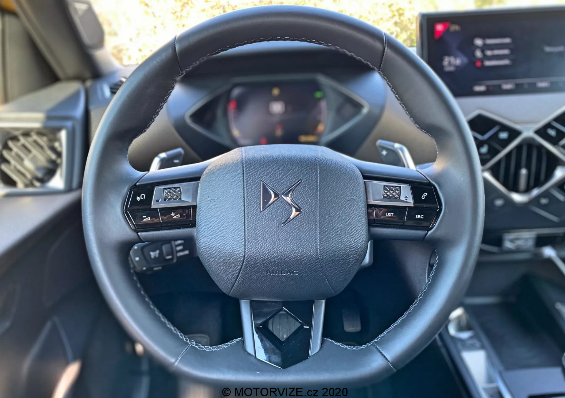 Záber interiéru automobilu DS Automobiles DS-3 Crossback z roku 2019 zameraný na luxusný kožený volant z pohľadu vodiča. Multifunkčný volant s ovládacími tlačidlami pre audio a informačný displej vozidla. Digitálny prístrojový panel a moderný dotykový informačný systém sú tiež čiastočne viditeľné, čo zdôrazňuje pokročilé elektronické rozhranie v tomto modernom automobilovom interiéri.