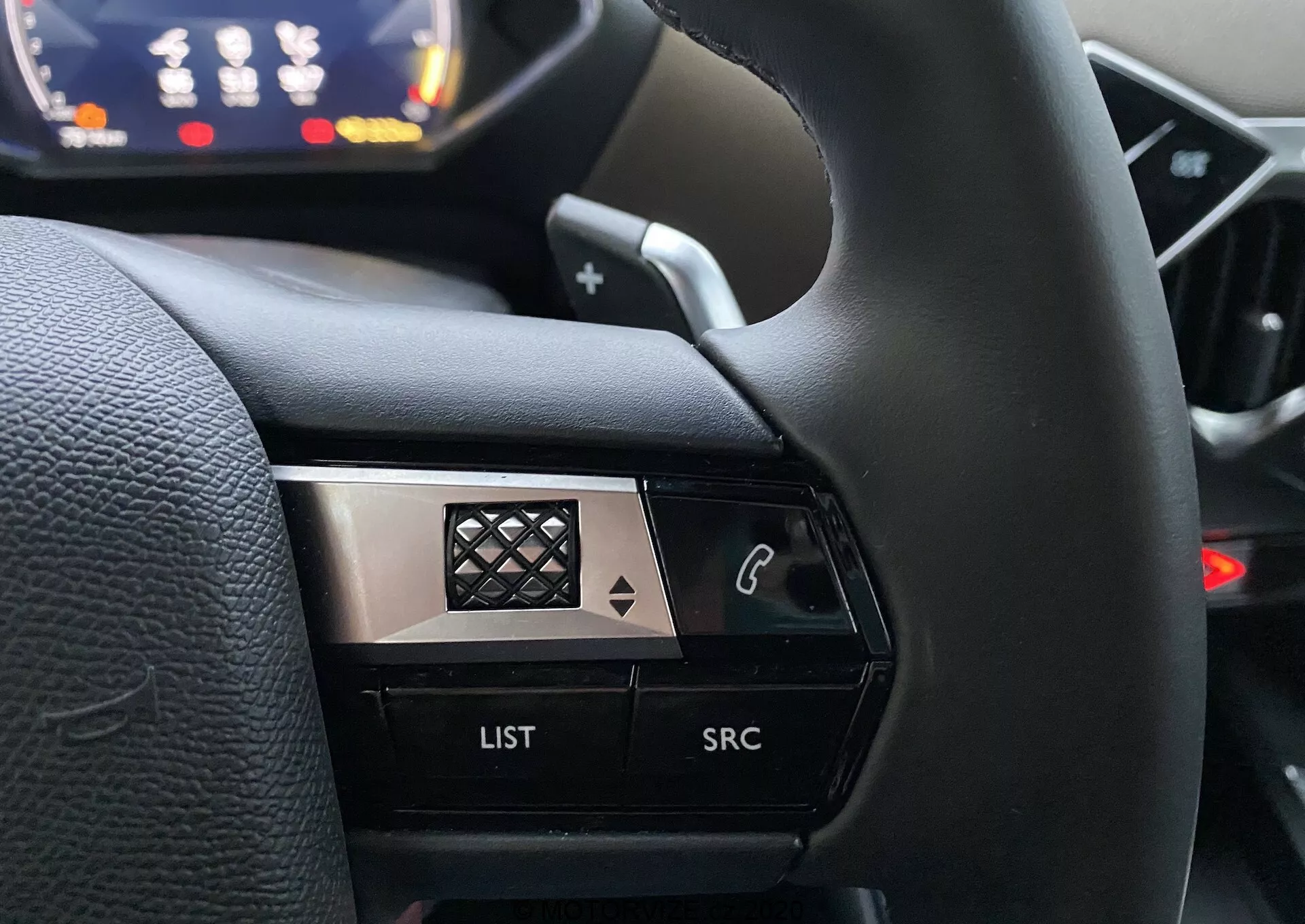 Detailný pohľad z perspektívy vodiča na ľavú stranu volantu v DS Automobiles DS 3 Crossback 2019. Obraz sa zameriava na ovládacie tlačidlá vrátane tlačidla s ikonou telefónu, tlačidla 'LIST' a tlačidla 'SRC', zvýraznené proti rozmazanému pozadiu palubnej dosky. Dizajn s vysokým kontrastom kombinuje čiernu a kovovú povrchovú úpravu s diamantovým vzorom na chróme, čo zvýrazňuje prémiový vzhľad.