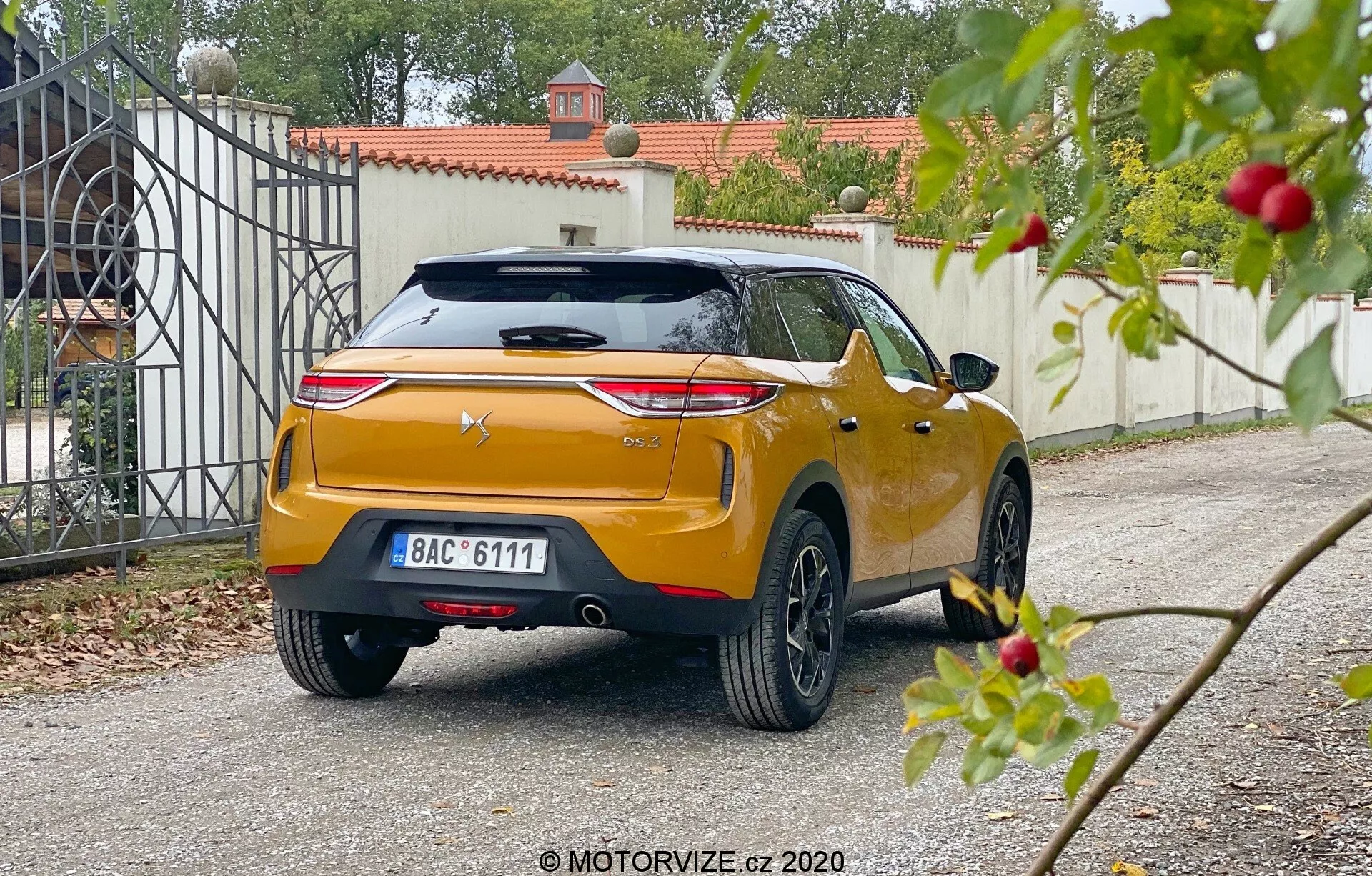 Zadný uhol pohľadu na DS Automobiles DS 3 Crossback 2019, zachytený z nízkej perspektívy na rezidenčnej príjazdovej ceste. Auto má výrazné horizontálne štýlové zadné svetlá, centralizované logo výrobcu na sochársky tvarovanom veku kufra s integrovaným spojlerom a tretím brzdovým svetlom, centrálne umiestnenú registračnú značku a lesklý metalický oranžový alebo zlatý povrch. Robustný zadný nárazník má integrované senzory alebo reflektory, viditeľný zadný stieračok, žraločí plochou anténu a viacpolohové zliatinové kolesá. Mierne rozmazaná zeleň v popredí kontrastuje s ostrejším zameraním na auto a vstupnou bránou na pozadí, čo naznačuje mäkké rozptýlené denné svetlo.